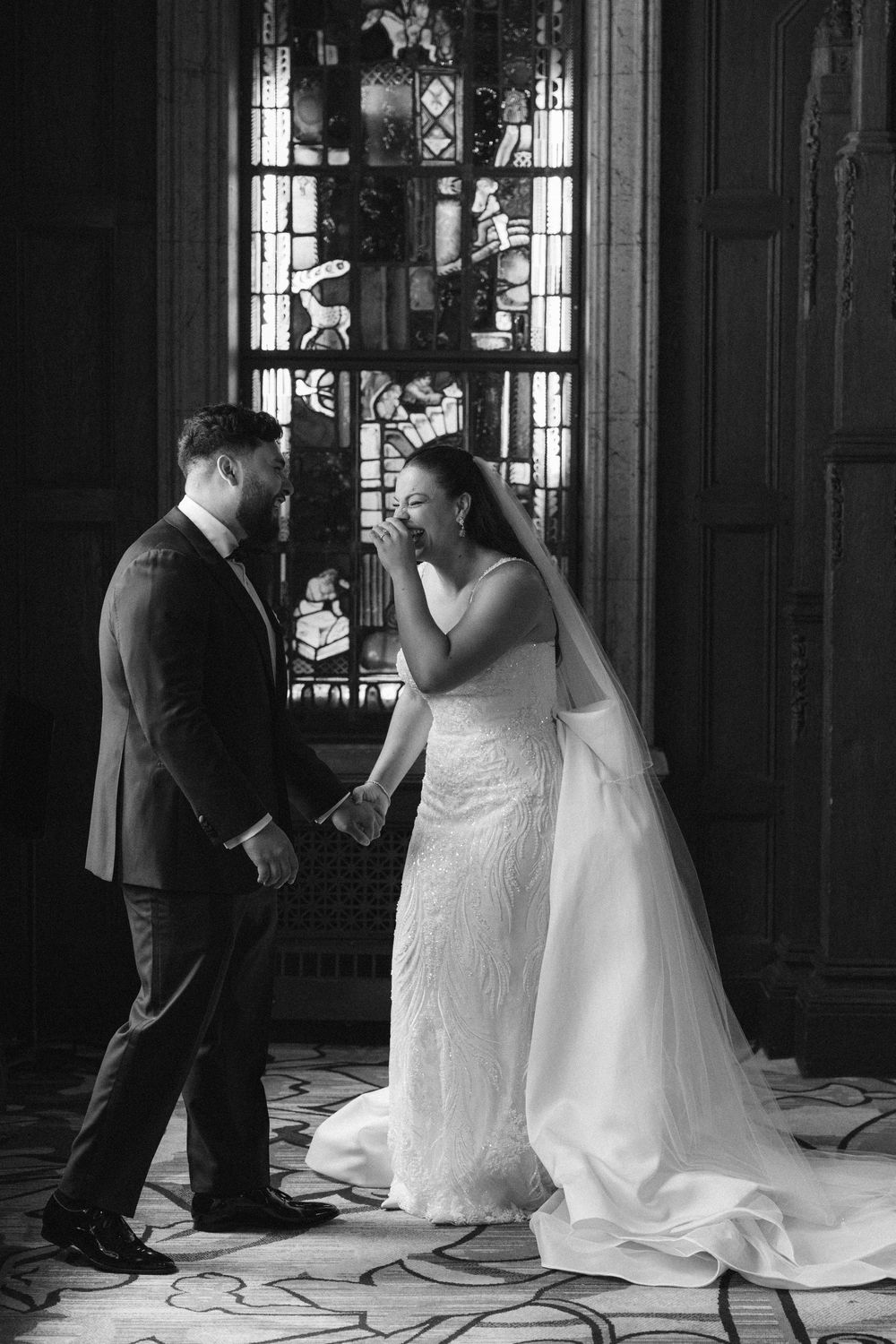 A black and white wedding photo of a couple sharing a candid moment in front of a stained glass window laughing and holding hands