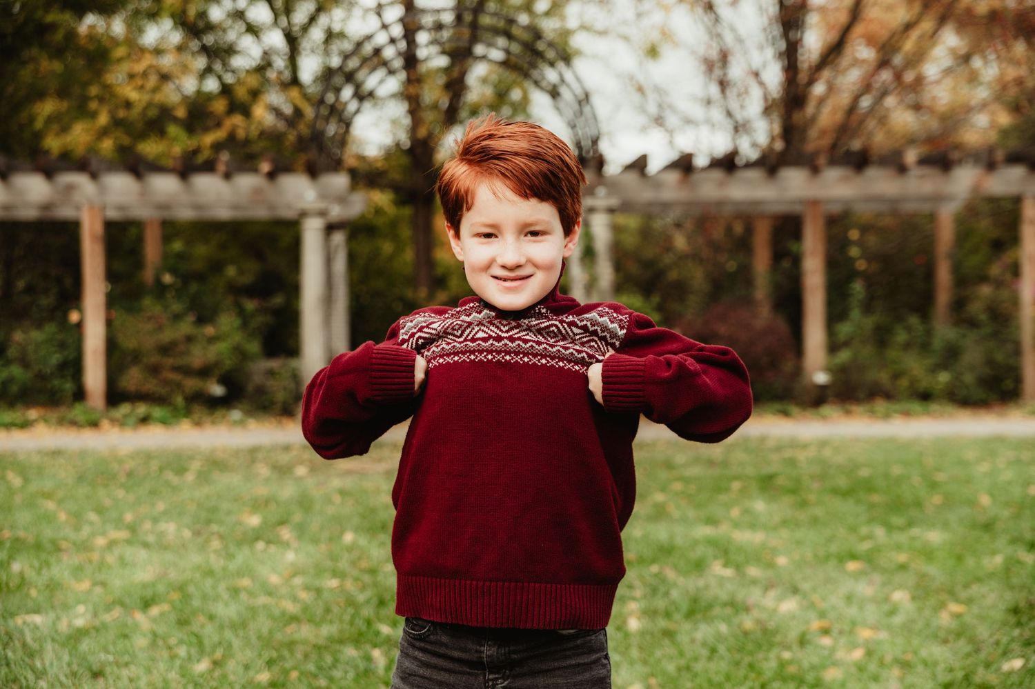Child wearing burgundy knit sweater poses playfully in garden setting.