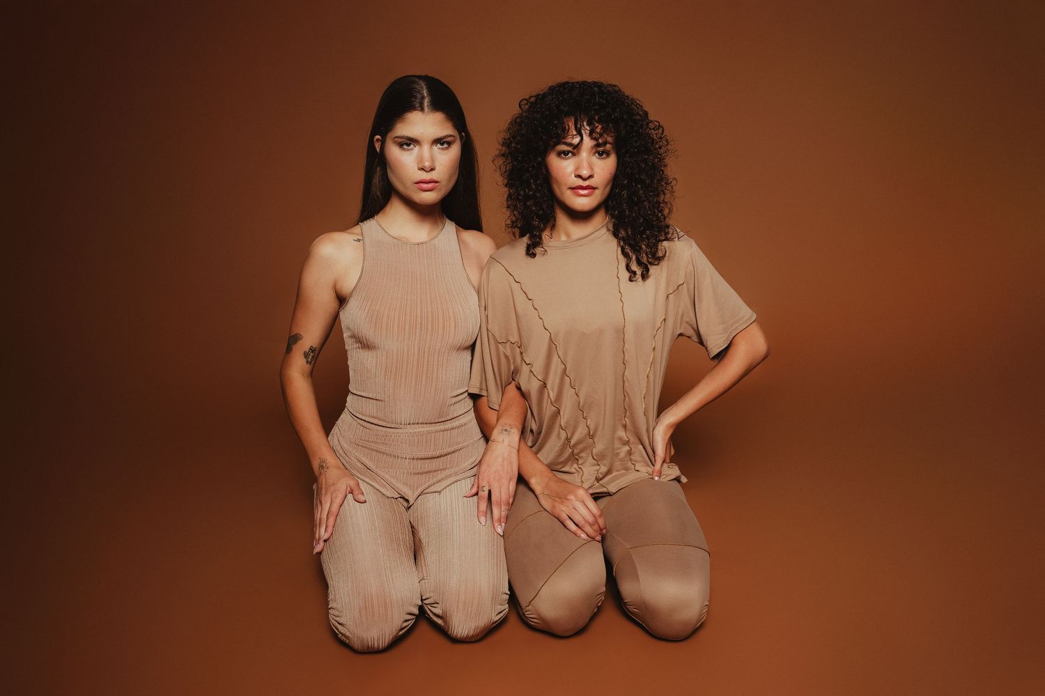 Models in neutral toned loungewear pose together against a warm brown backdrop showing different clothing styles.