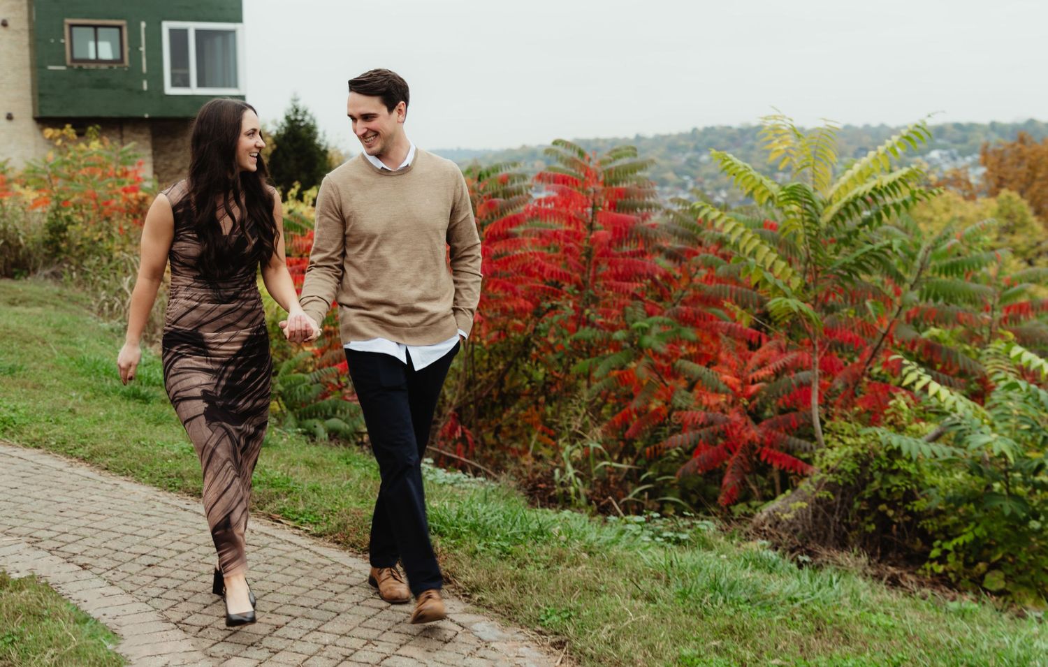 A couple strolls along a garden path lined with vibrant red autumn foliage and bushes.