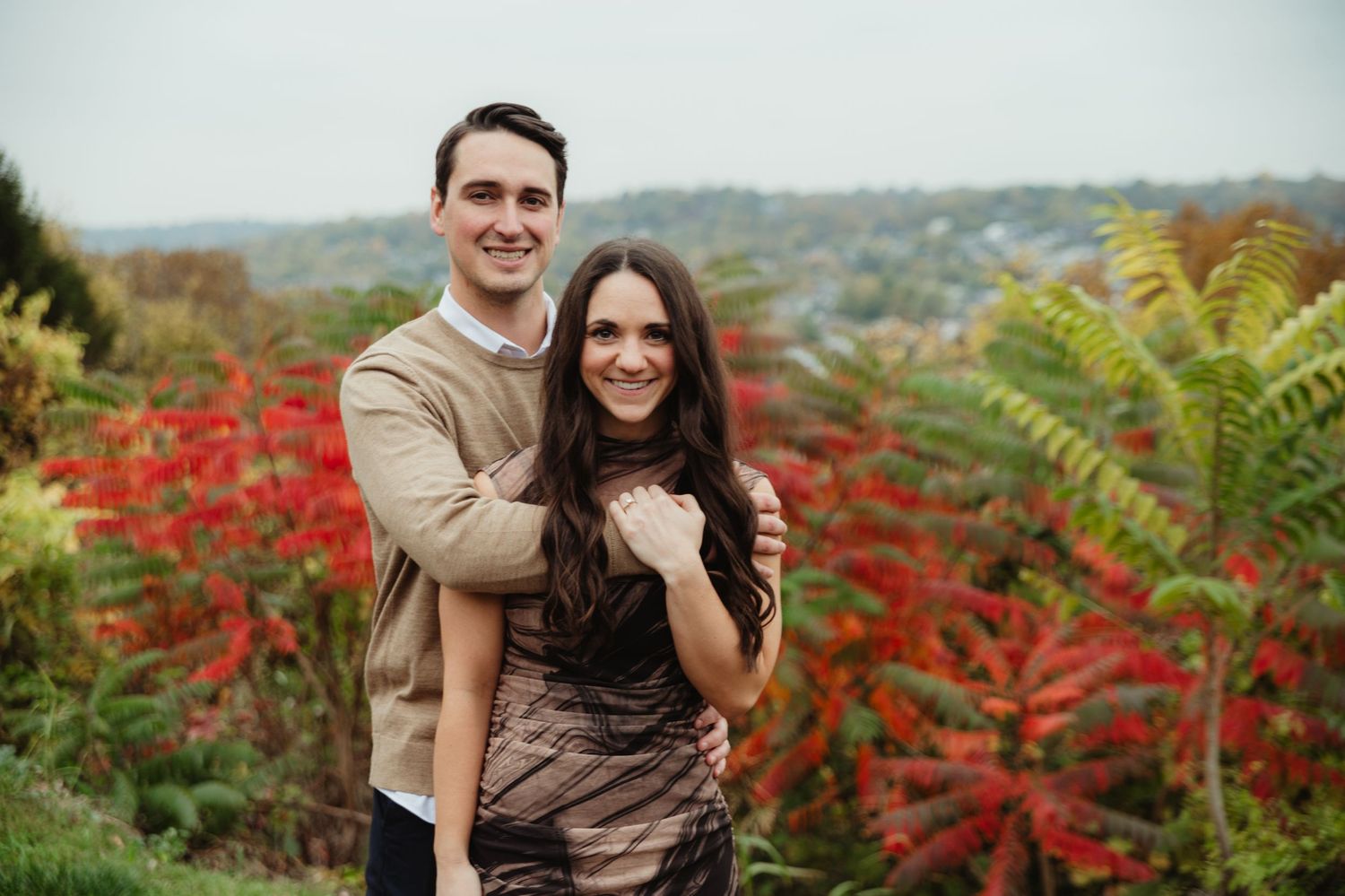 A couple poses for engagement photos in a scenic autumn landscape with red foliage and ferns.