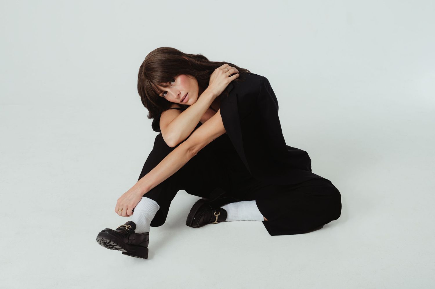 Person in black clothing crouches on floor in artistic dance pose against light background.