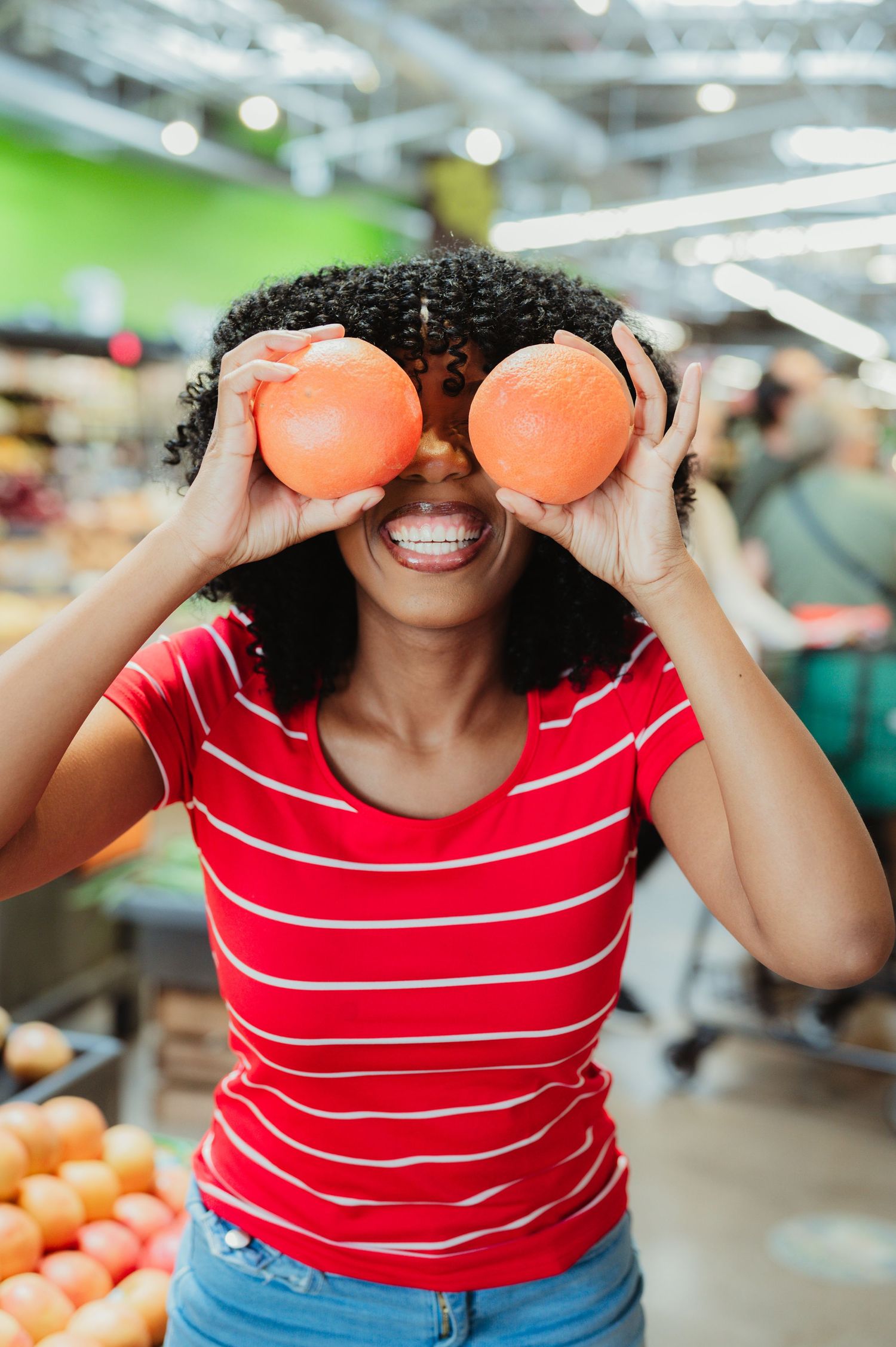 Someone in a red striped shirt playfully holds oranges over their eyes while shopping at a produce market.