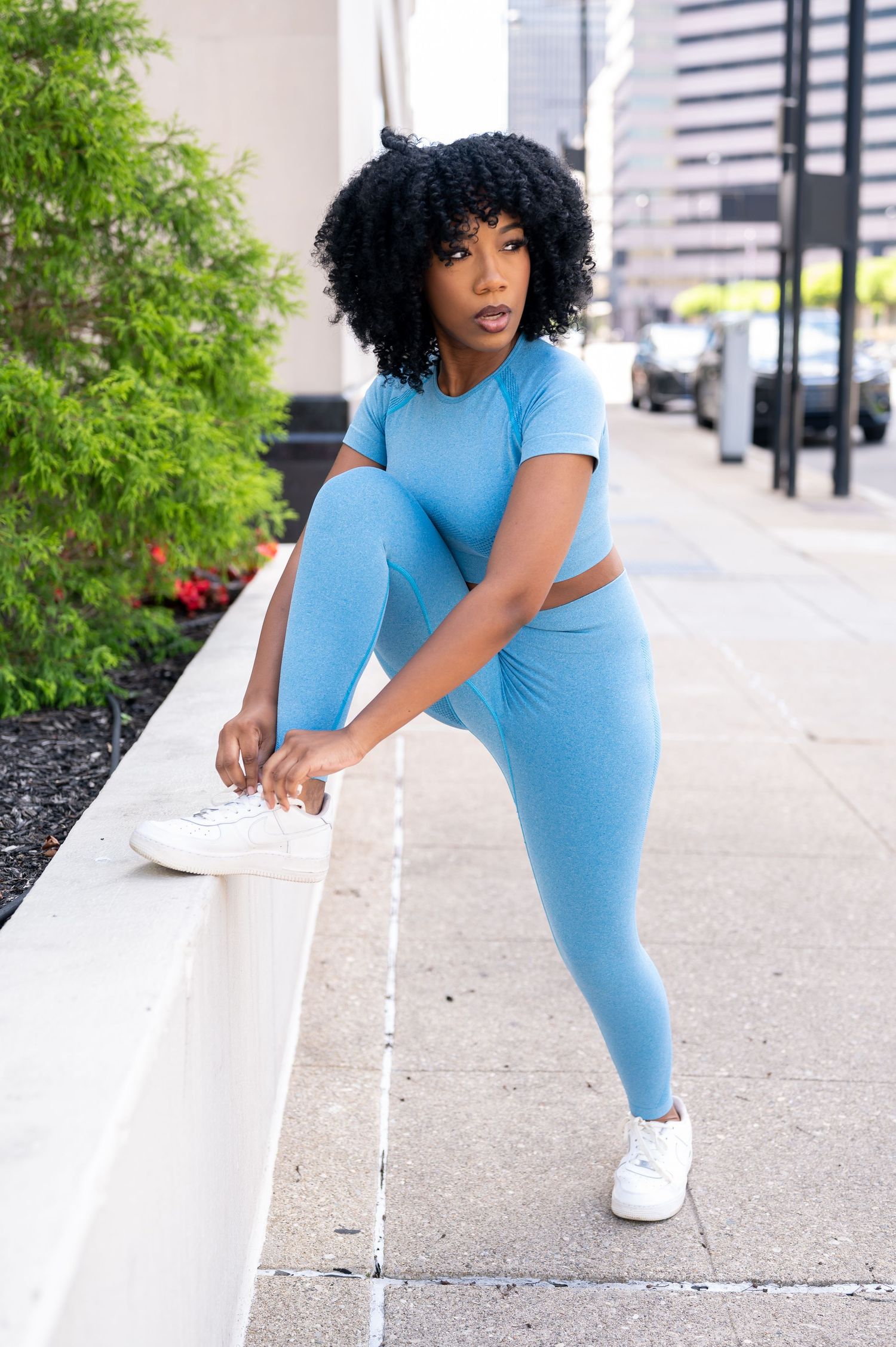 Athletic figure in light blue workout set and white sneakers stretches outdoors in an urban setting.