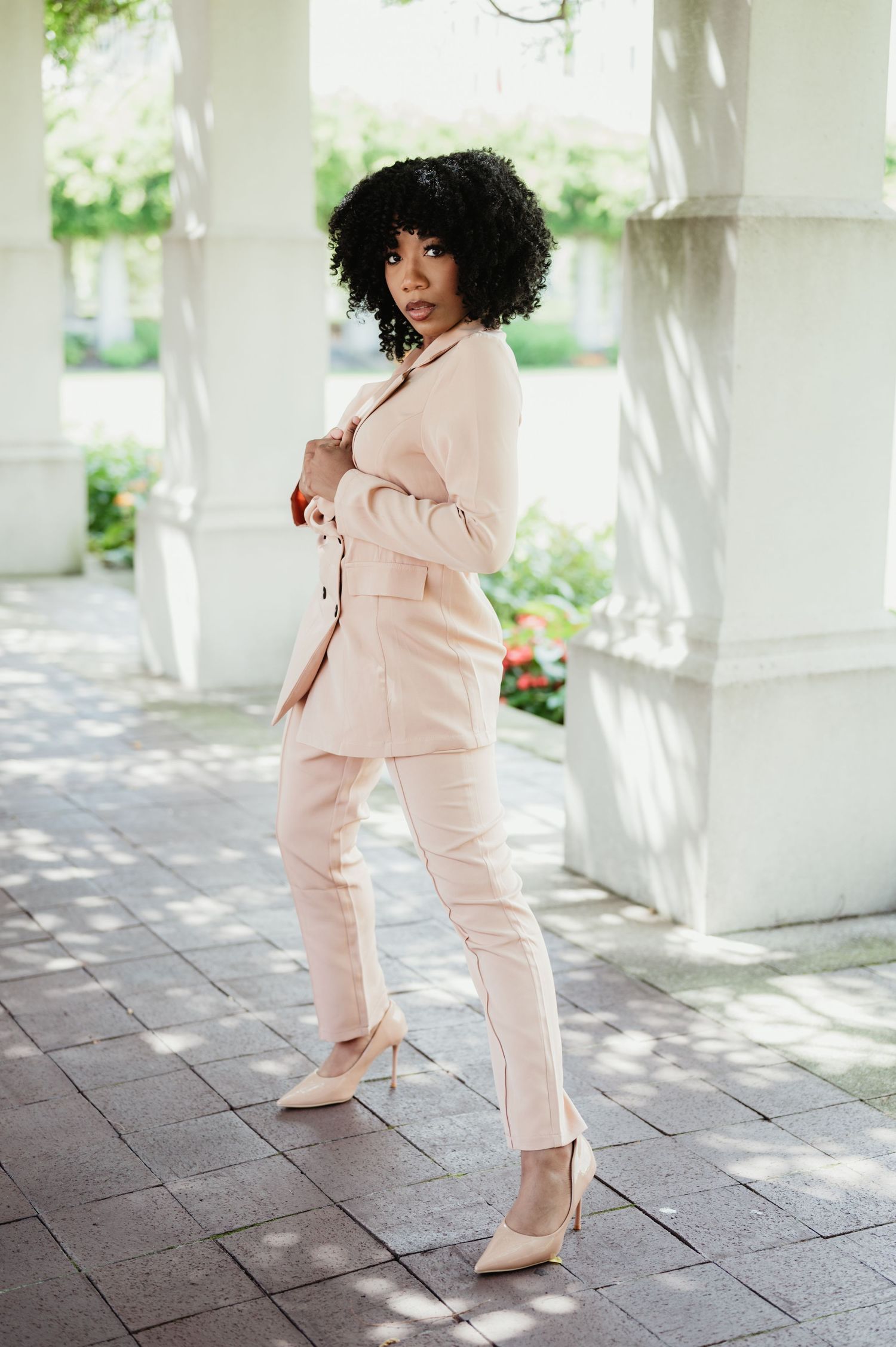 A person in a soft pink tracksuit poses elegantly on a paved walkway surrounded by white columns and greenery.