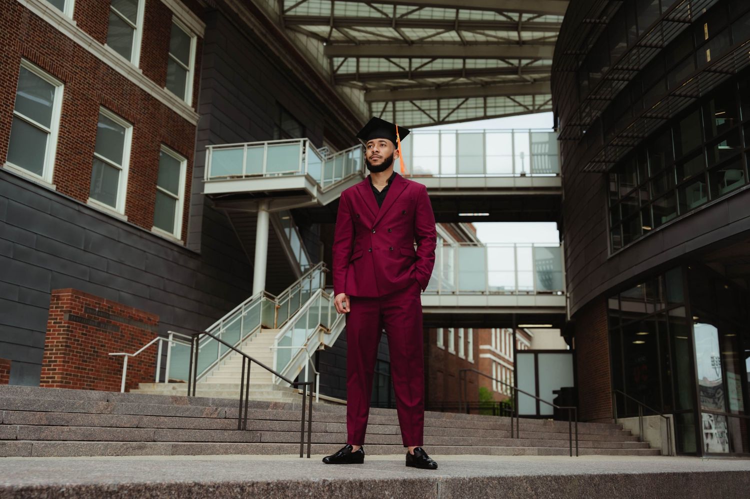 A person in a burgundy suit stands confidently on concrete steps beneath an industrial-style skybridge.