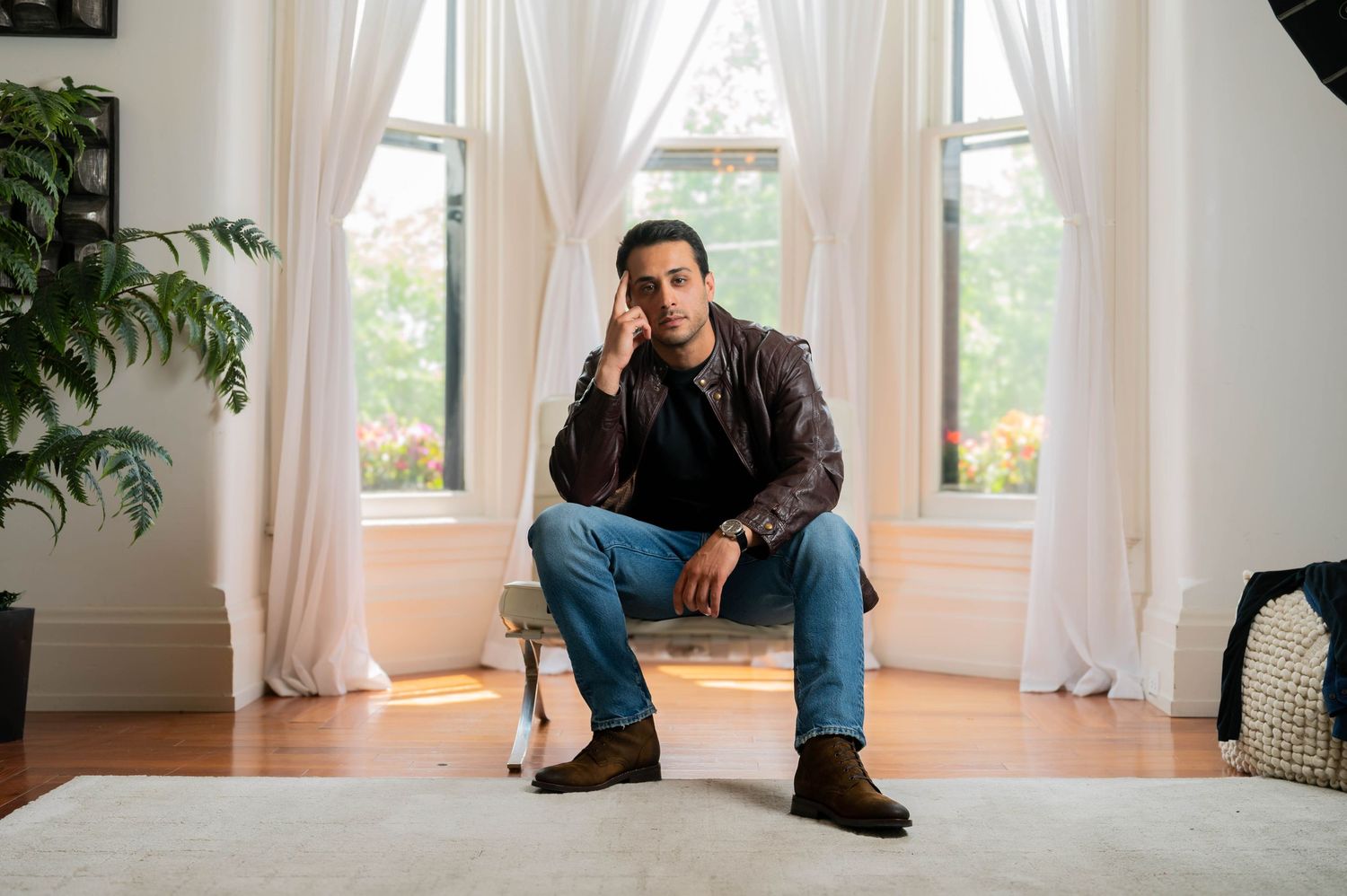 A person sits on a windowsill in a bright room with white curtains and hardwood floors.