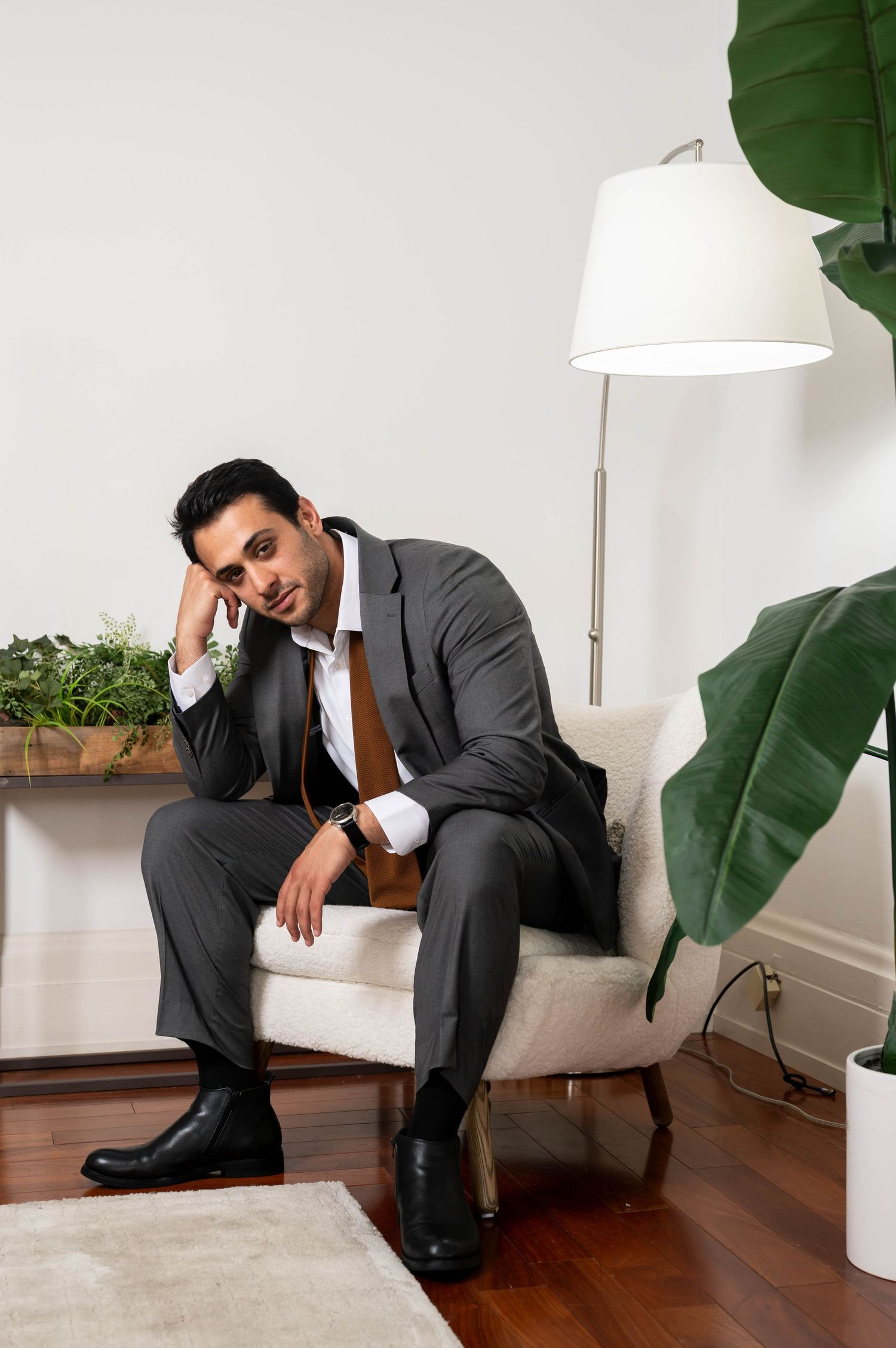 Person in grey suit lounges on white sofa with large green plants in modern minimalist interior setting.