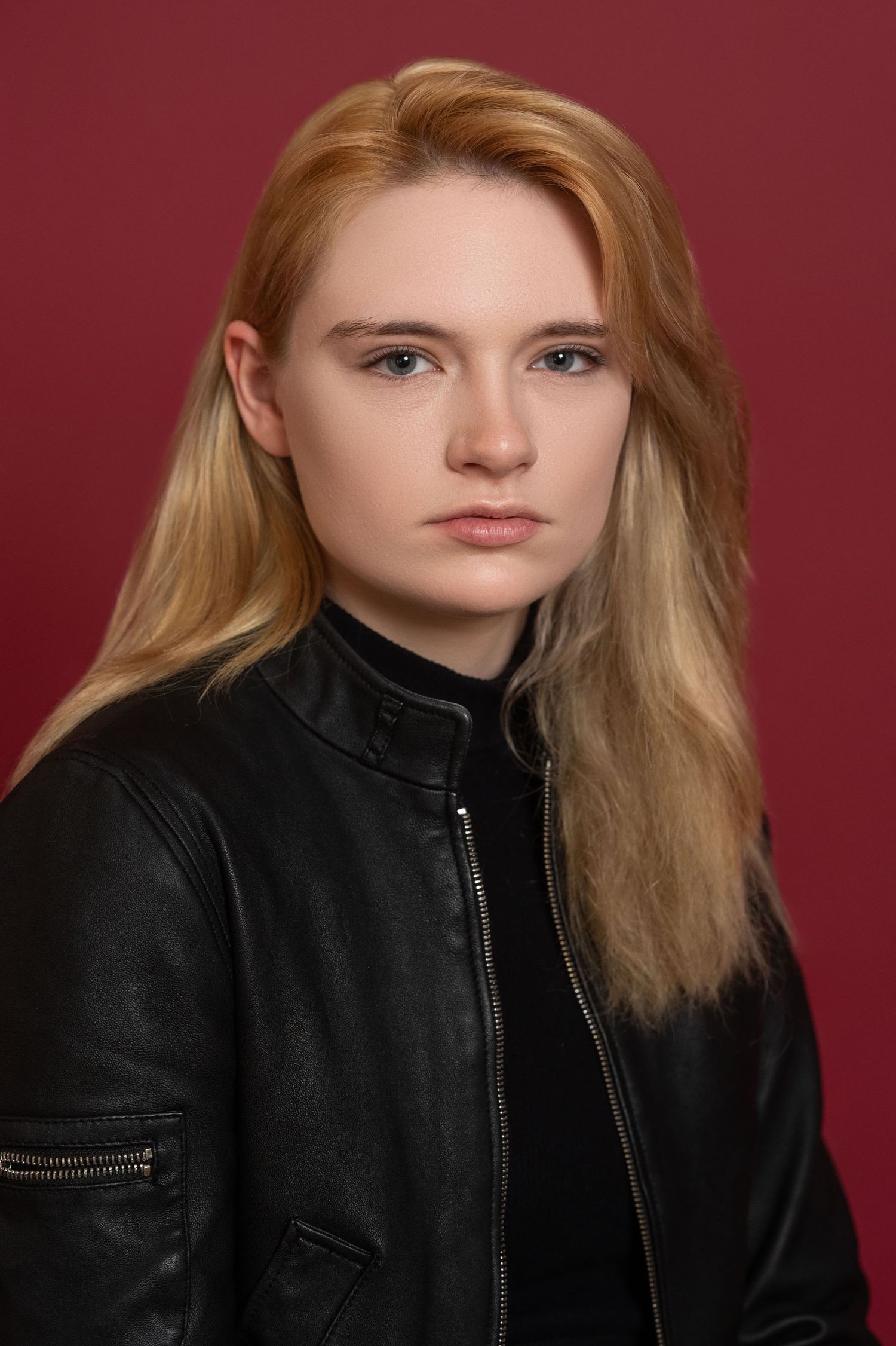 Portrait against burgundy background featuring blonde hair and black leather jacket in dramatic lighting.