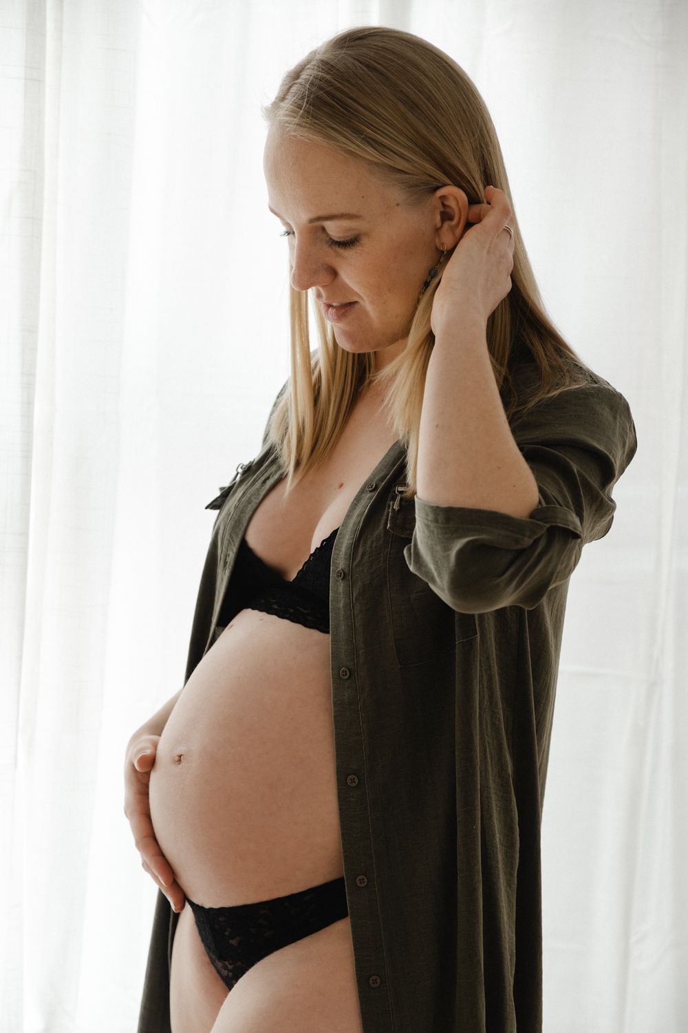 A pregnant woman in black lingerie and an olive green button-down shirt stands by a window in soft natural light.