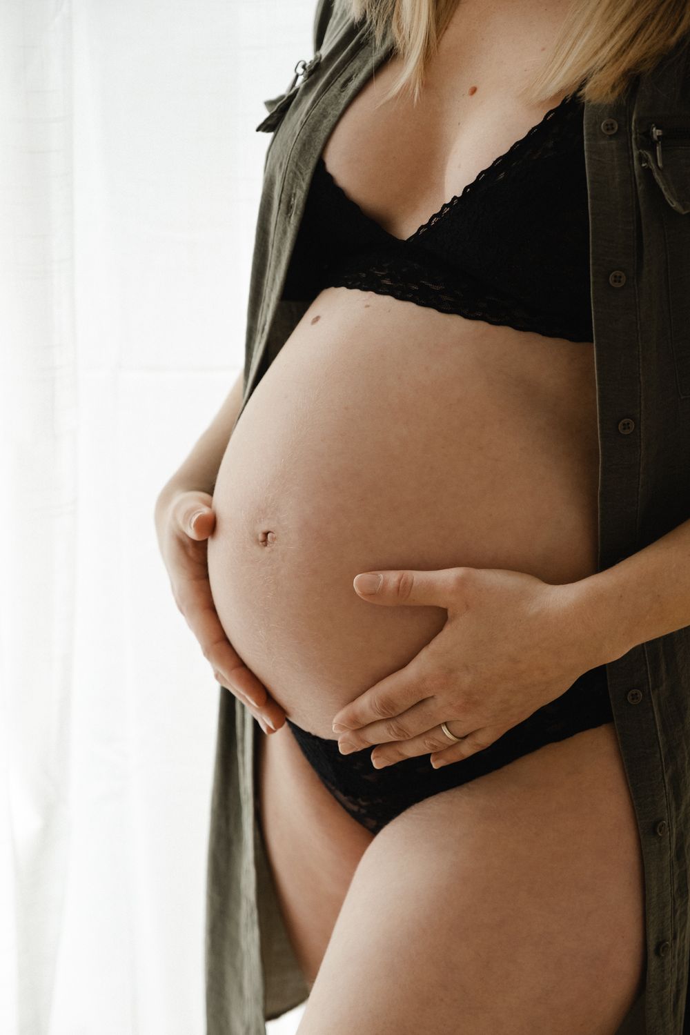 Artistic maternity photo of a pregnant belly with hands cradling bump, wearing black lingerie and olive green button down.