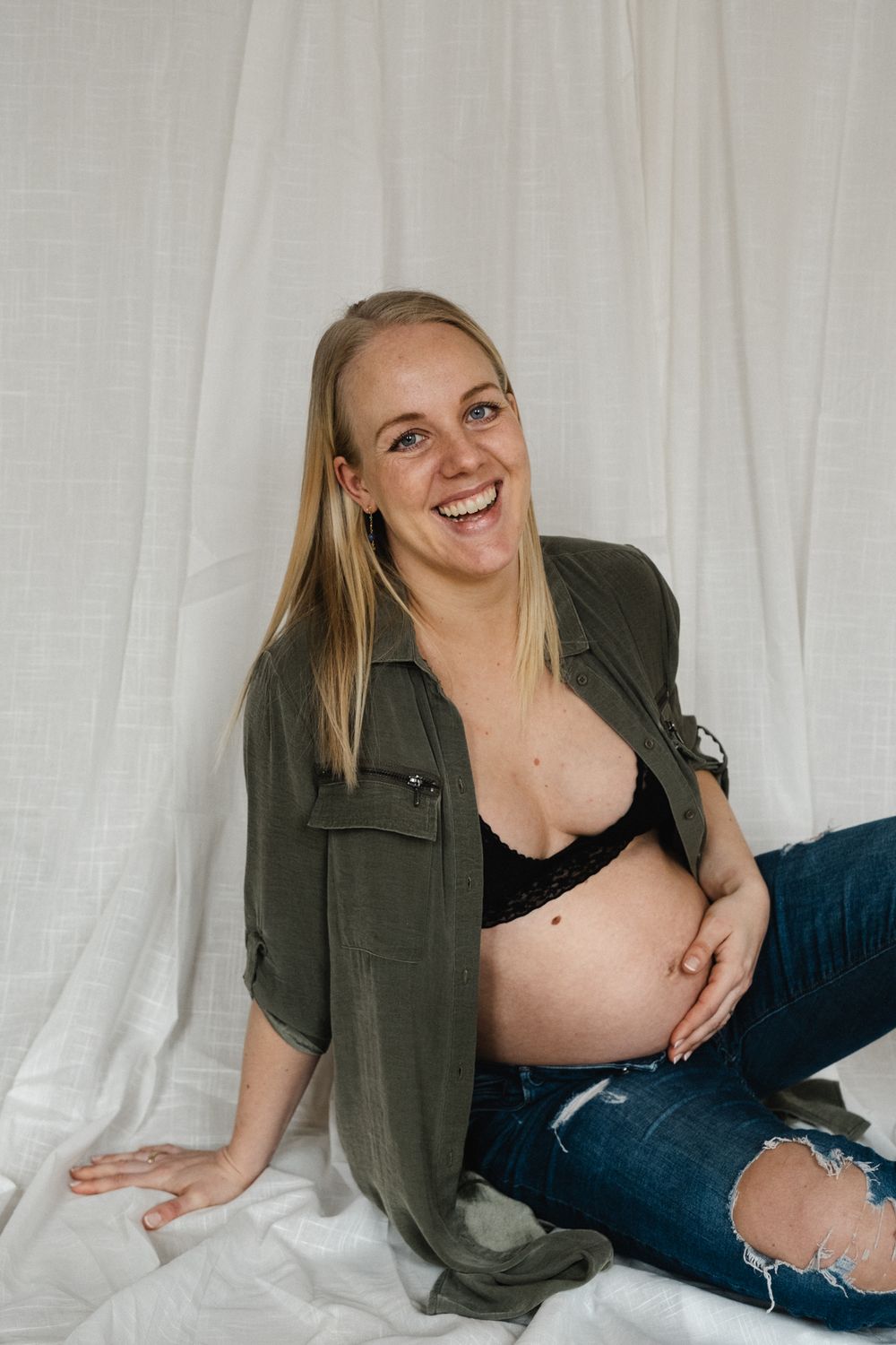 Maternity photo of pregnant woman wearing olive green shirt and distressed jeans against white backdrop.