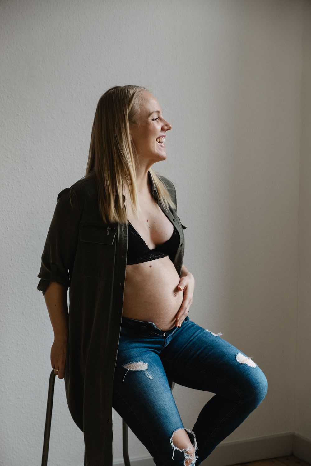 Side profile of an expectant mother in distressed jeans and open shirt laughing while posing against a light wall.