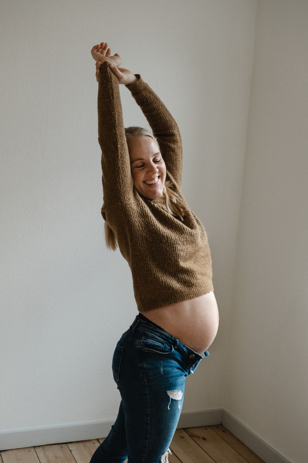 A pregnant person stretches joyfully in a brown sweater and jeans while standing against a white wall.