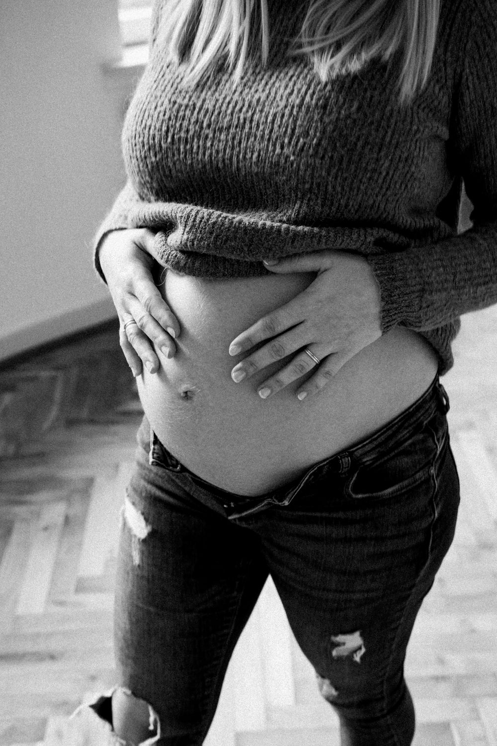 Black and white photo of a pregnant belly in a knit sweater and distressed jeans showing maternity silhouette.