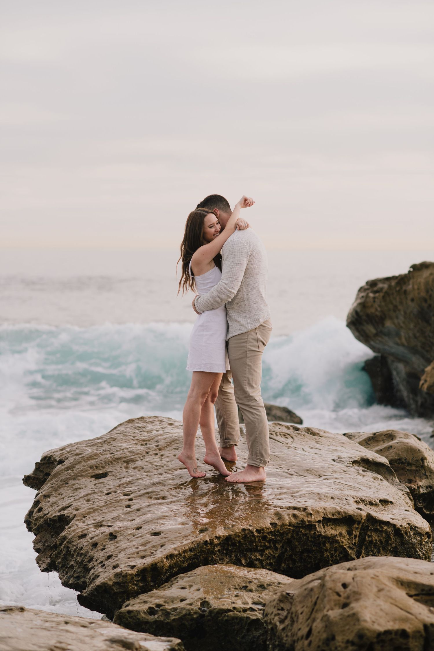 Barefoot couple embraces on rocky shoreline with dramatic ocean waves crashing in the background.