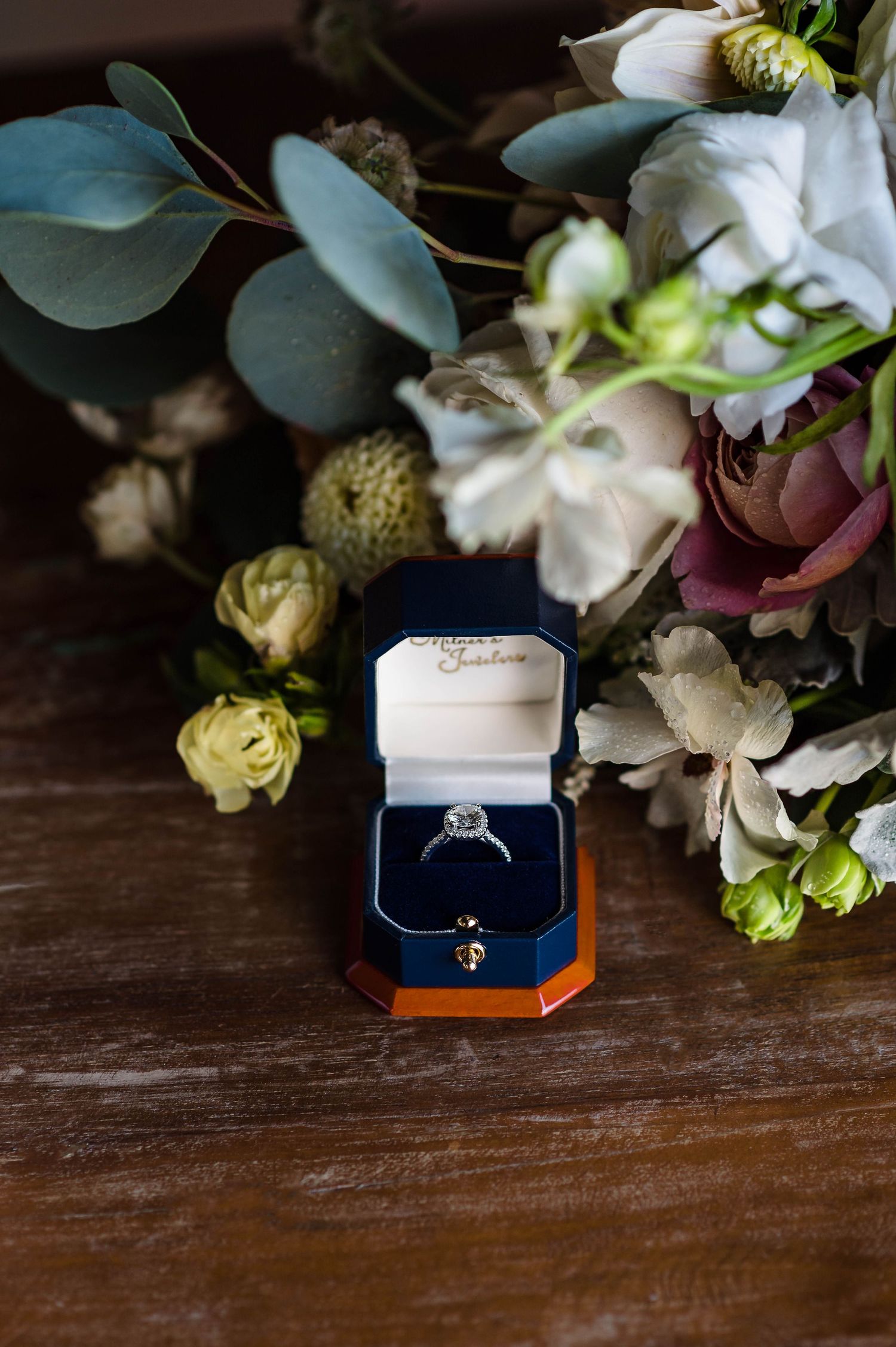 Engagement ring in a navy blue velvet box displayed with white and green floral arrangements on a dark wooden surface.