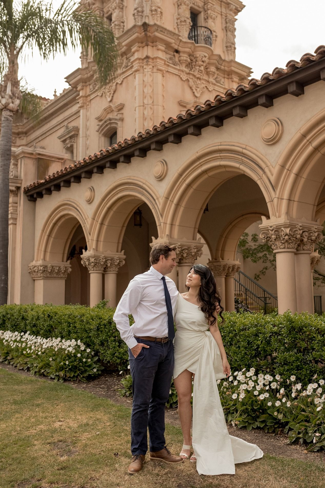 A couple shares romantic moments in front of a Spanish colonial style building with arched walkways and white flowers at Balboa Park.