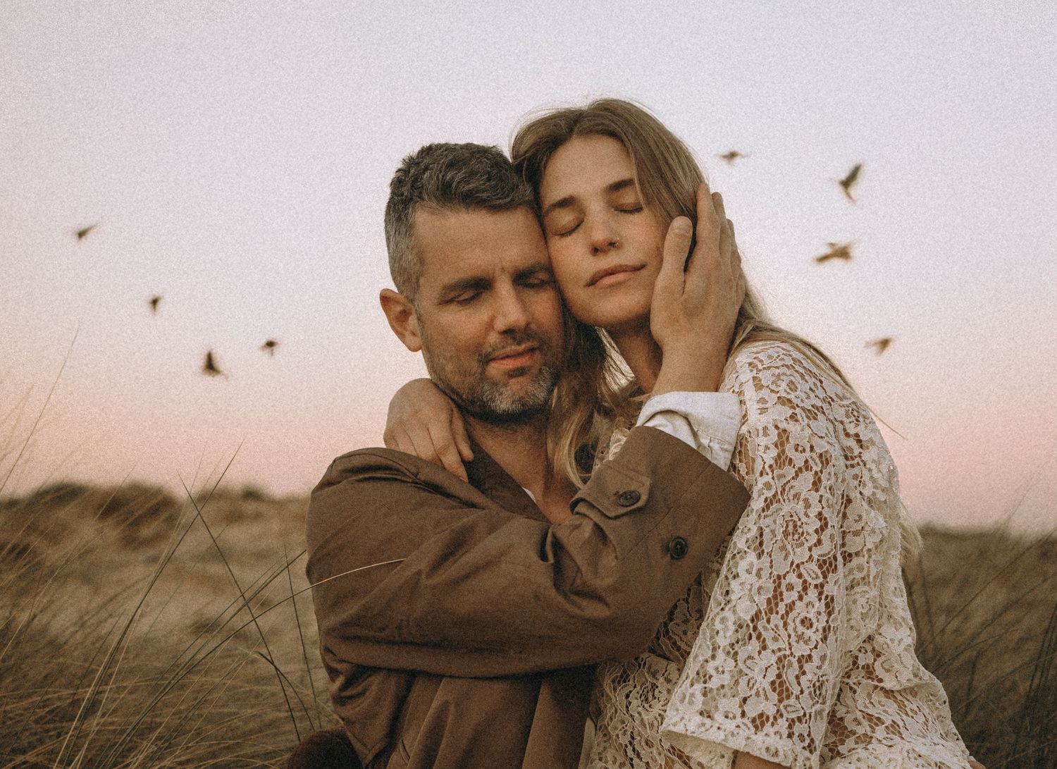 A couple in rustic attire embraces tenderly in a field at sunset with butterflies floating around them.