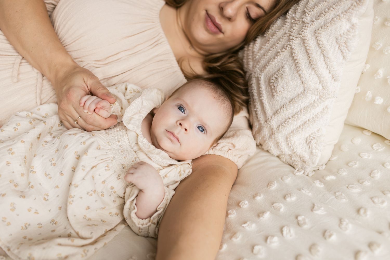 Newborn girl wearing a cream floral dress snuggled into the crook of her mother's arm while laying on a cream colored bedspread in Andover.