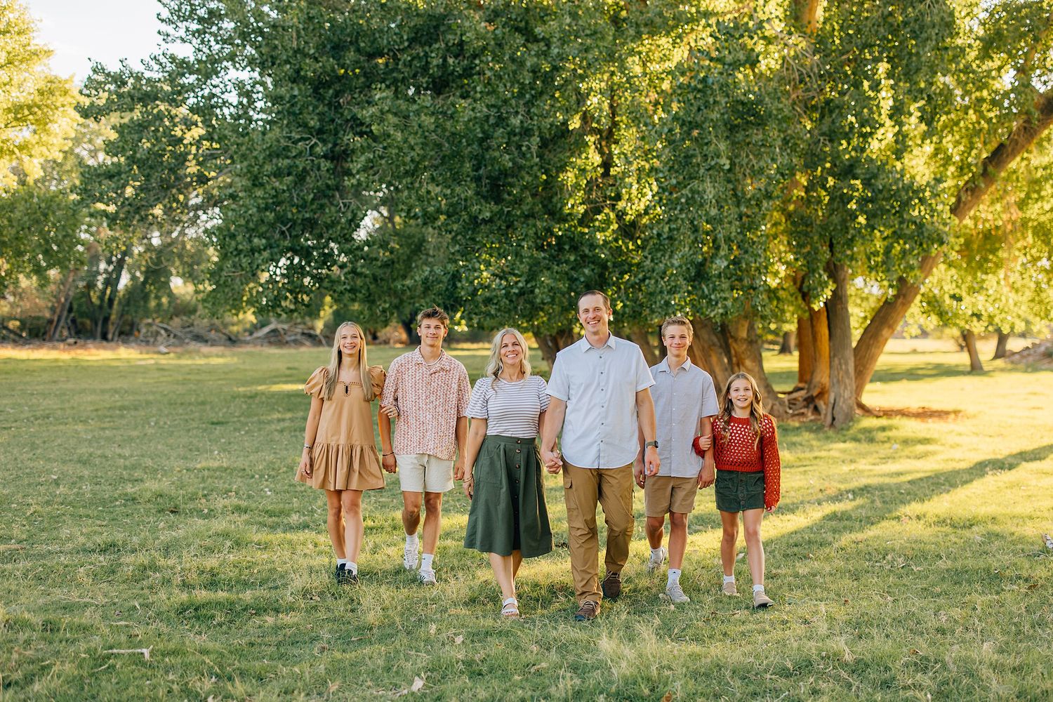 A group of people in casual summer attire walk together across a sunlit grassy field with tall trees in the background.
