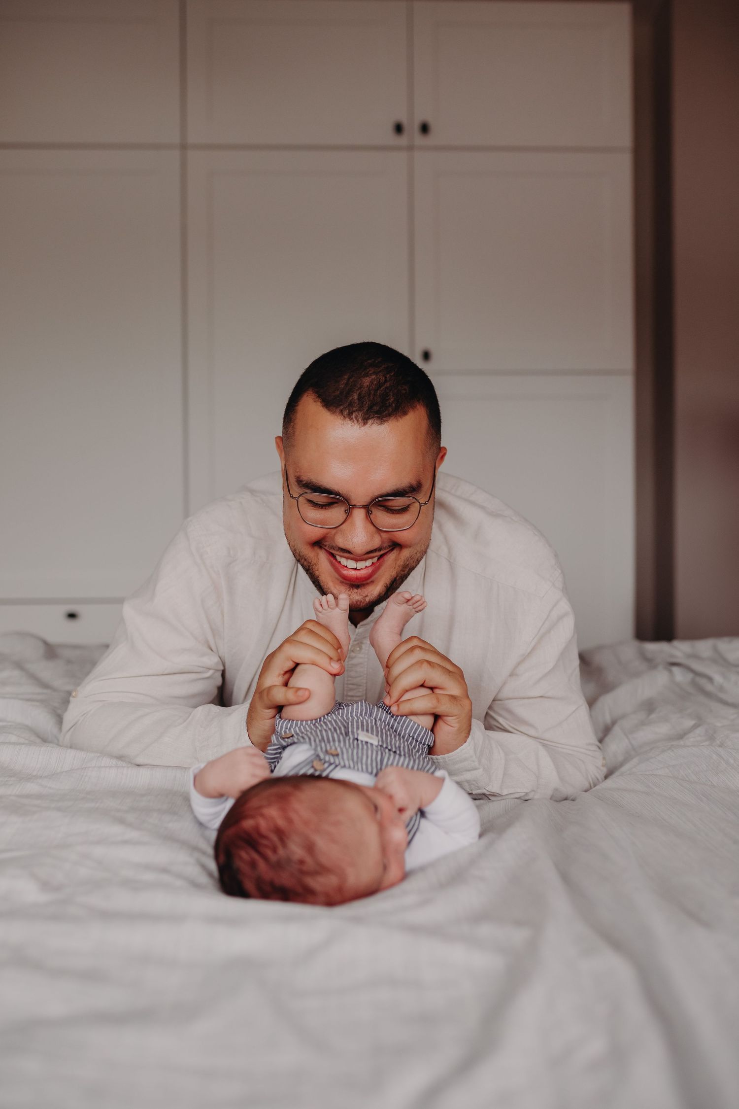 Smiling father with glasses playing with newborn baby on white bed at home.