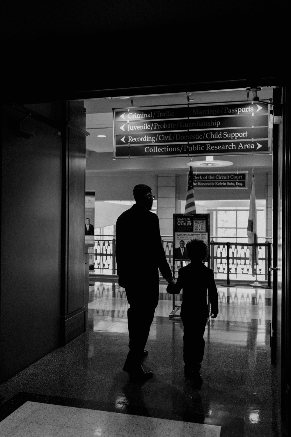 A silhouetted adult and child walk together through an airport terminal hallway under directional signage.
