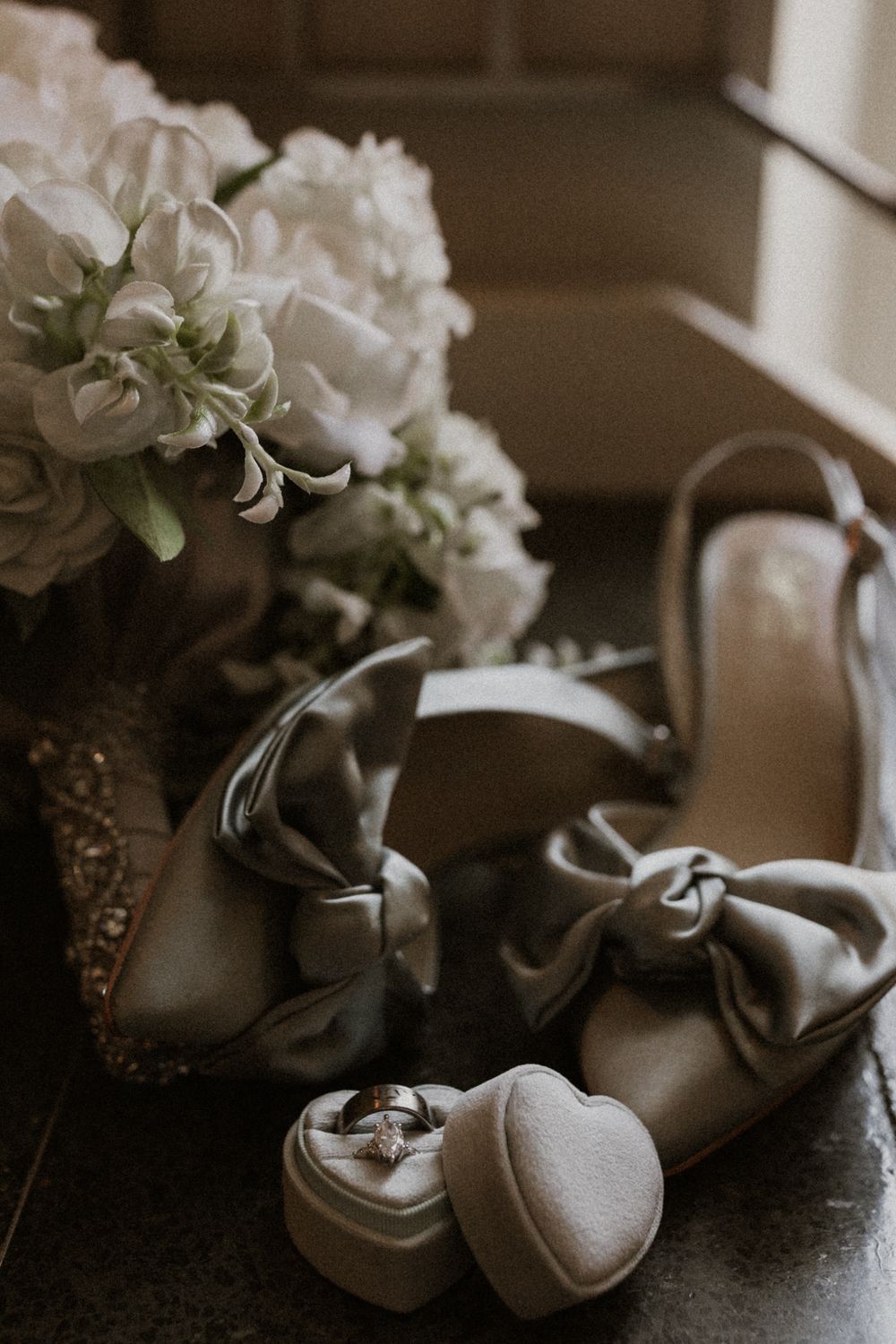 Wedding details featuring elegant satin shoes with bows alongside white floral bouquet in moody lighting.