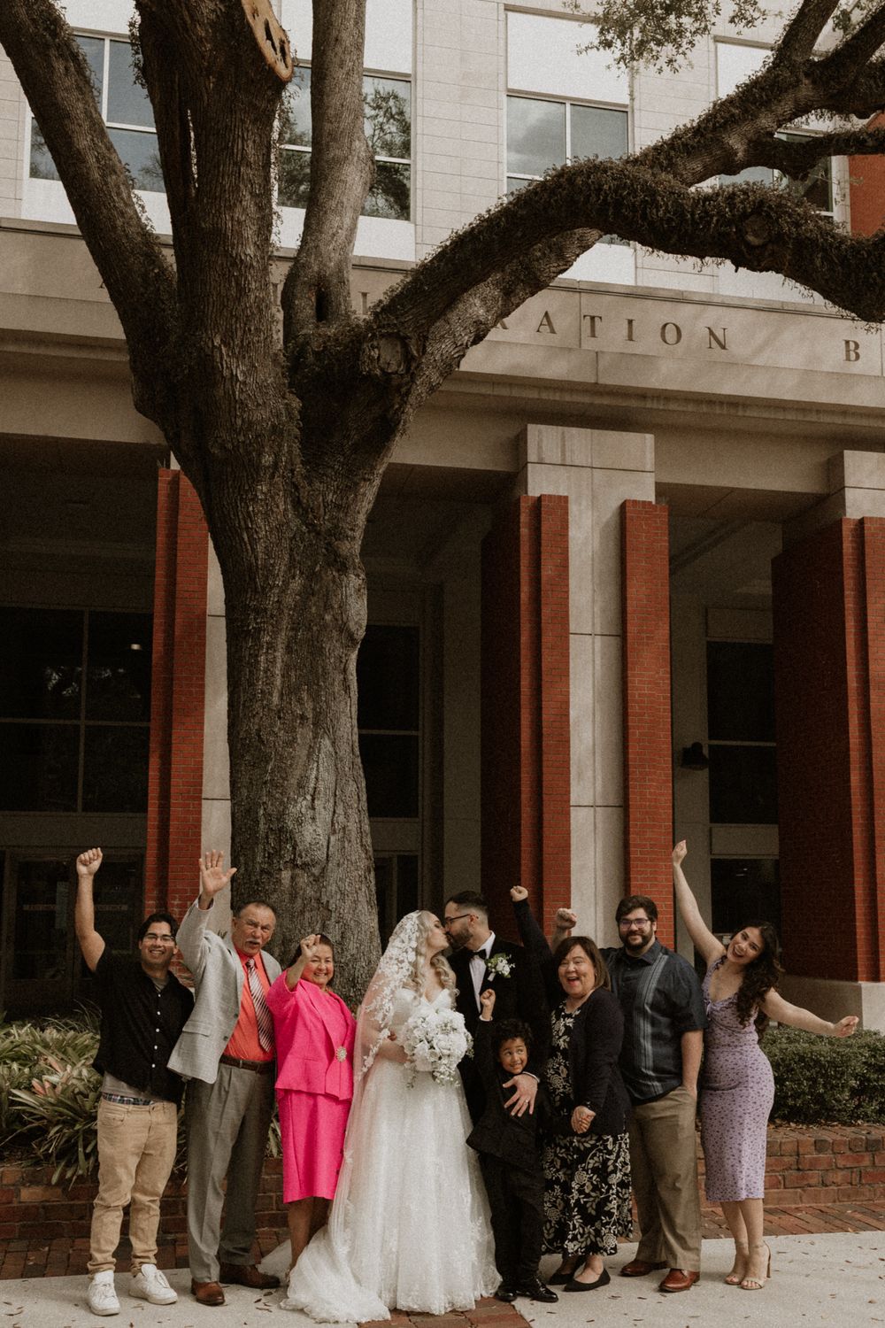 Wedding party poses playfully behind trees outside a brick and stone building.