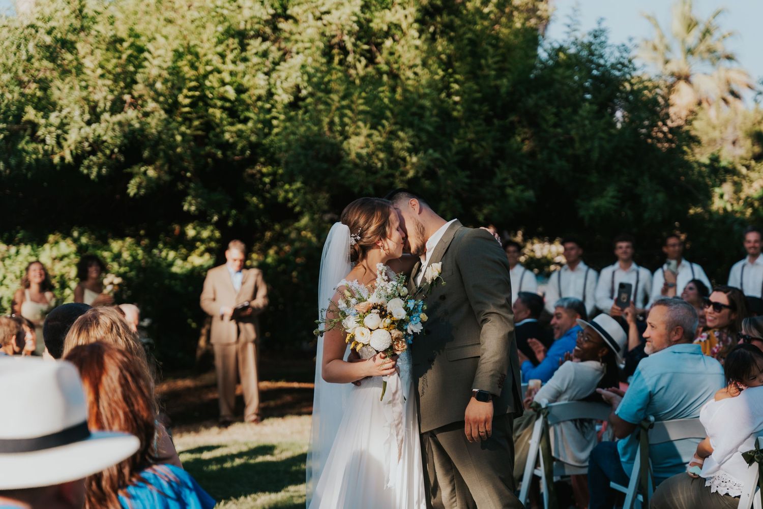 A couple shares a kiss during an outdoor wedding ceremony surrounded by guests and lush greenery on a sunny day.