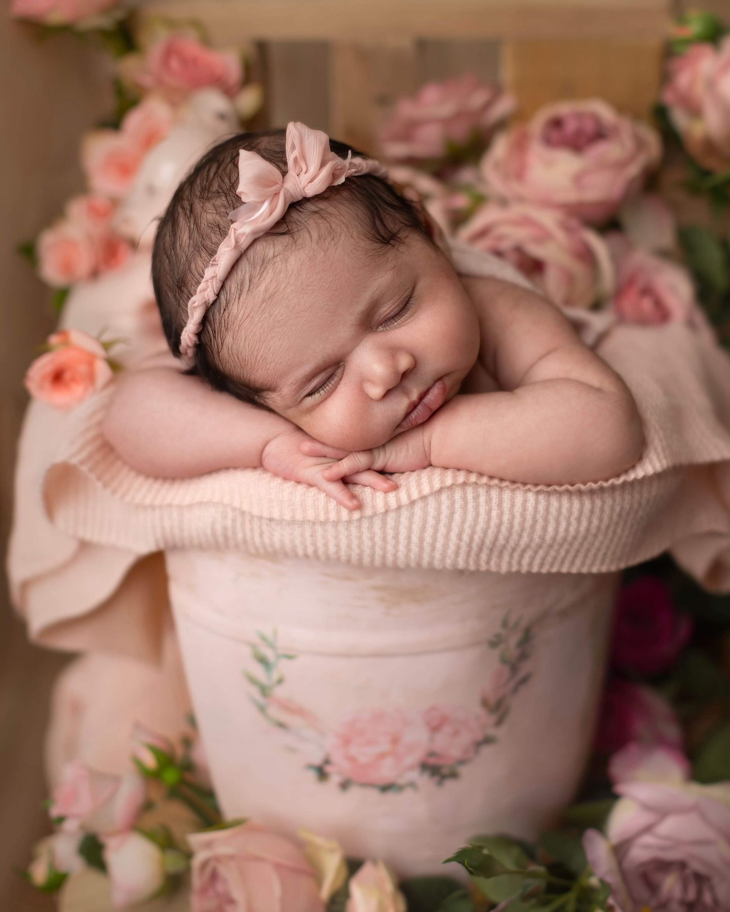 A sleeping newborn nestled in a pink floral teacup surrounded by delicate roses and soft lighting. Elizabeth Lang Photography Surrey