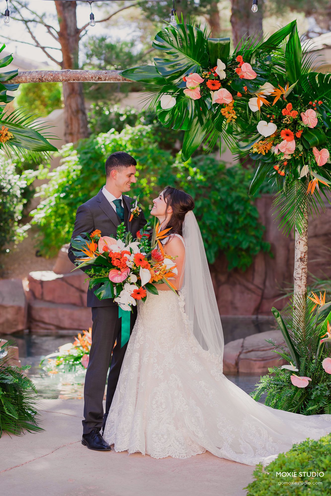 Wedding couple poses under tropical floral arch with coral roses and palm leaves in lush garden setting.