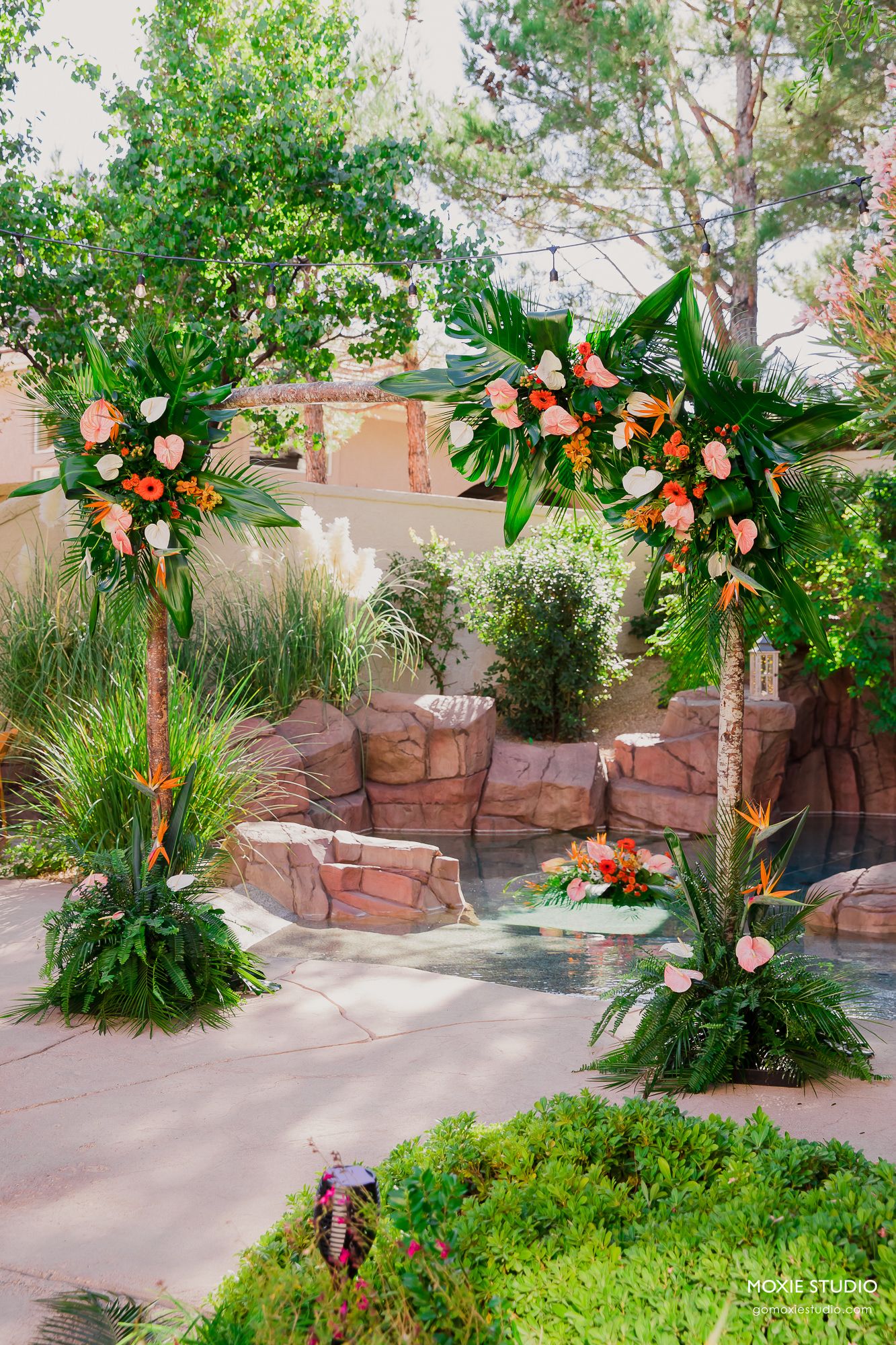Tropical floral arch with peach roses and greenery decorates a stone garden patio setting for an outdoor ceremony.