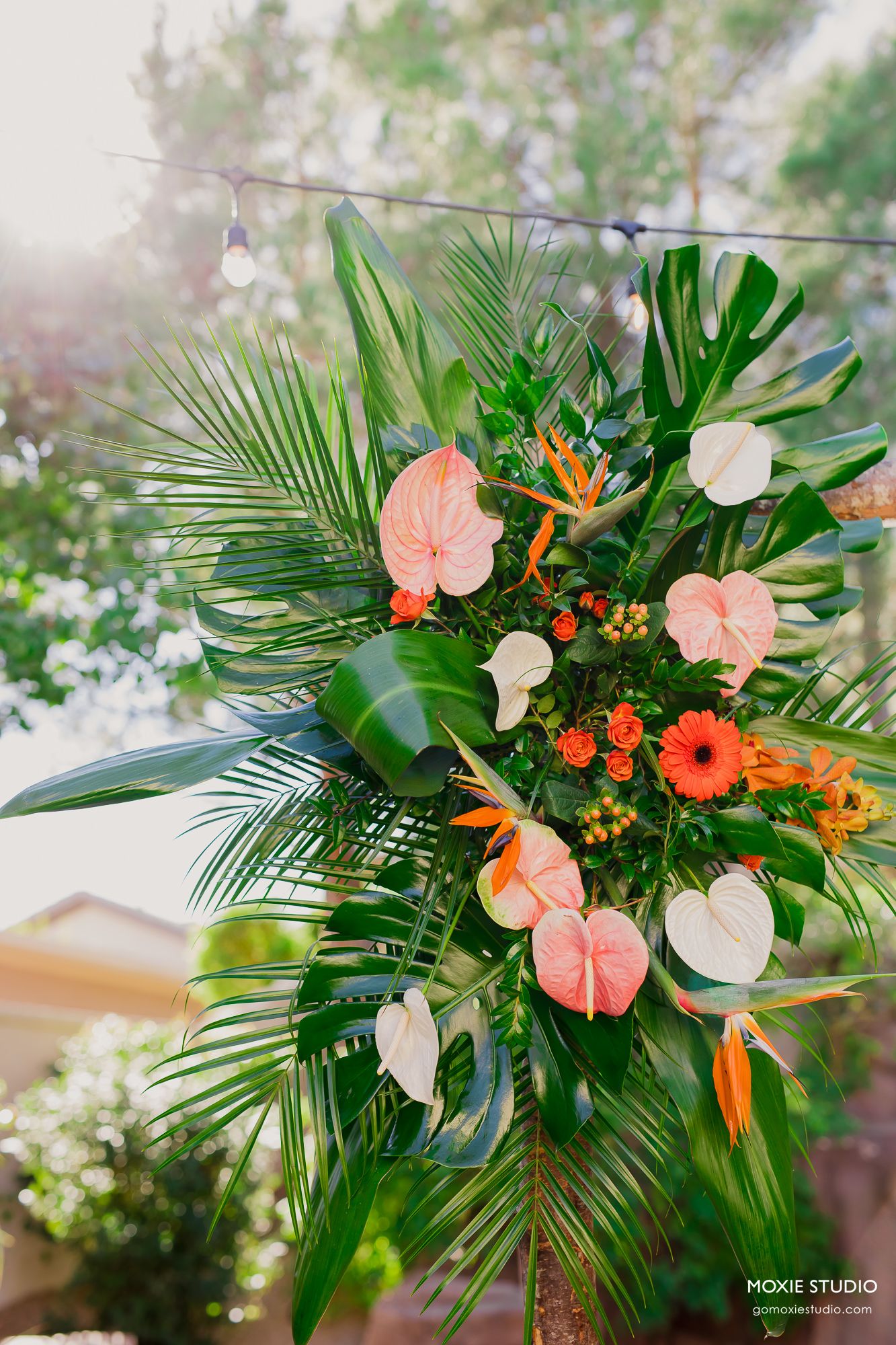 Tropical floral arrangement with orange gerbera daisies, pink anthurium, and white flowers among palm fronds and greenery.