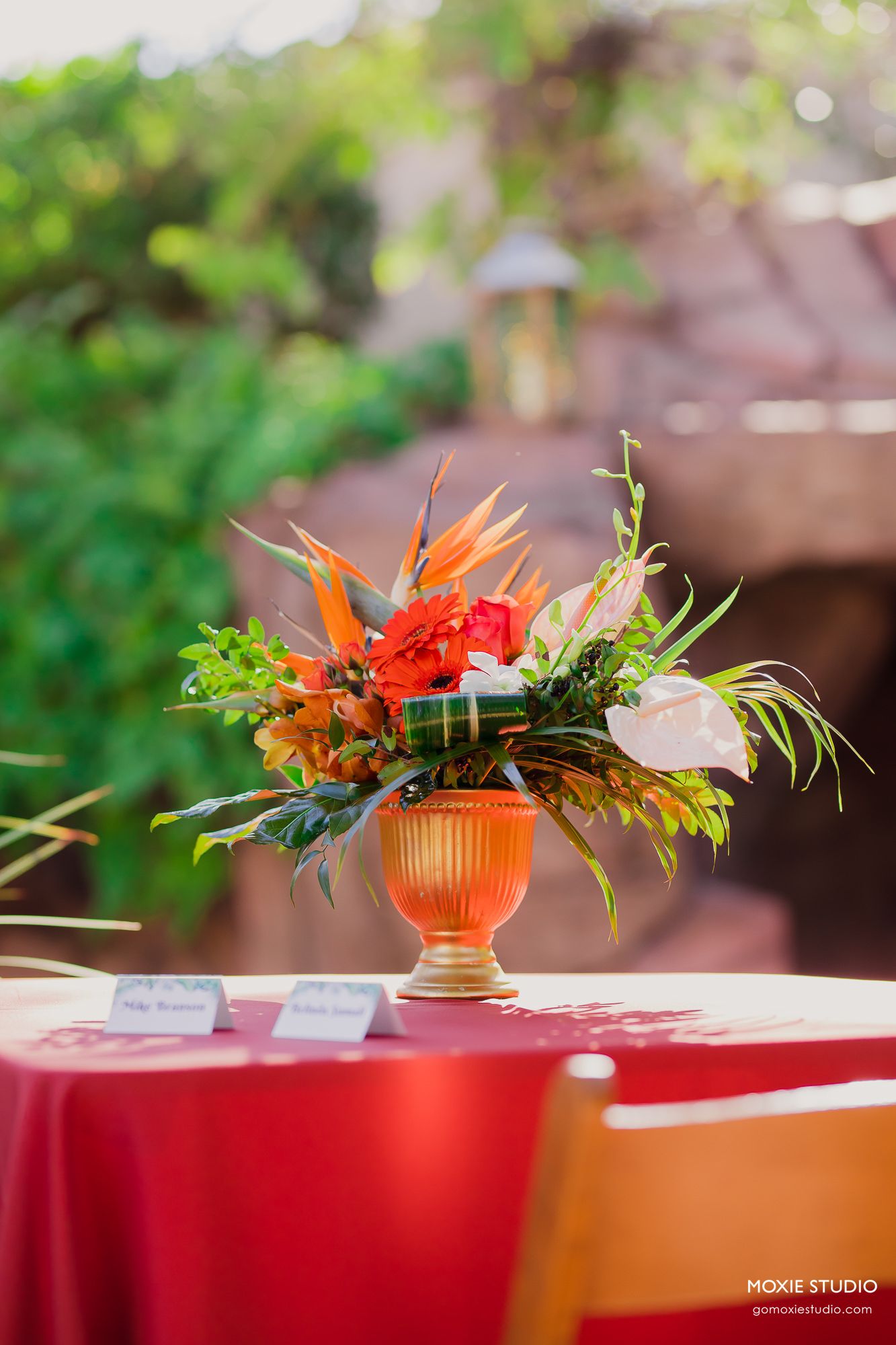Vibrant orange and red tropical flowers arranged in an amber glass vase on a red tablecloth at an outdoor event.