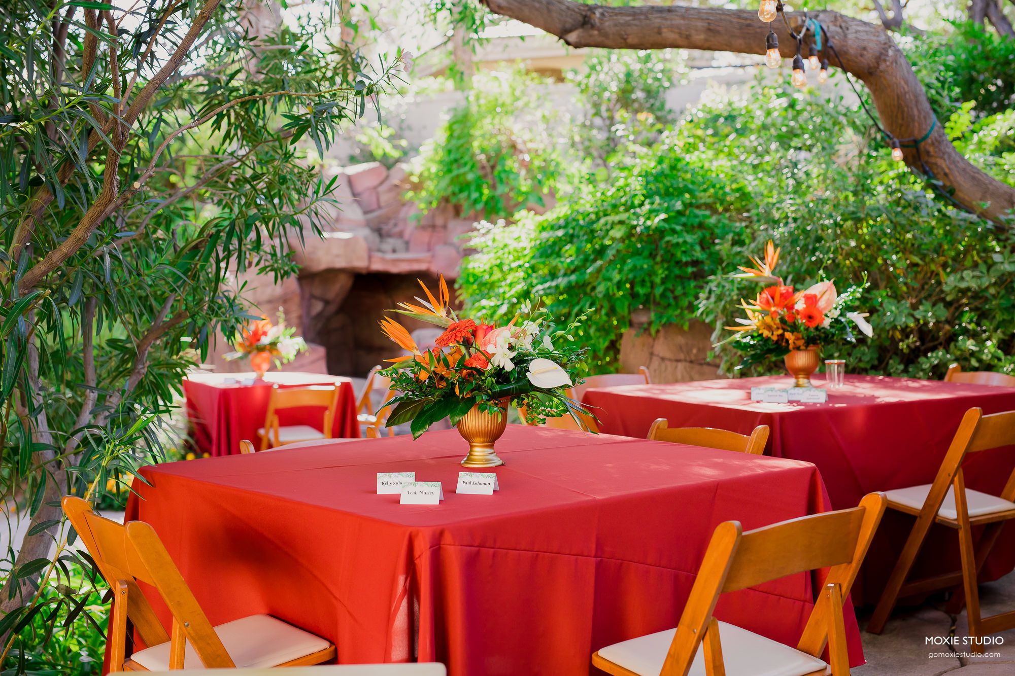 Red tablecloths and wooden chairs create an elegant outdoor dining atmosphere under lush green trees.