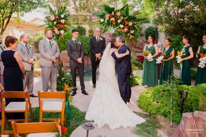 A wedding ceremony takes place in a lush garden setting with floral arrangements and attendants in green dresses.