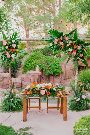 Tropical floral arrangements with palm fronds and coral roses decorate wooden benches in an outdoor garden setting.