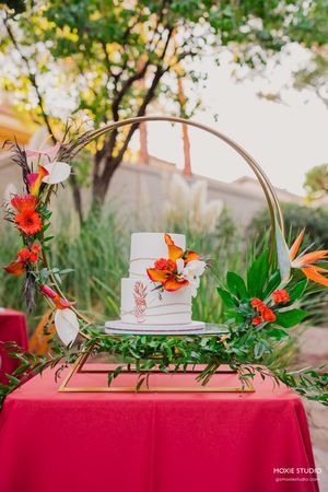 Elegant wedding cake display on red tablecloth with tropical flowers and greenery decorations in outdoor garden setting.