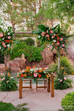 Tropical floral arrangements with orange birds of paradise and pink roses adorn wooden benches in an outdoor garden setting.