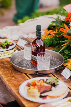 A bottle of red liquor on a serving tray with crystal glasses alongside plates of food on a rustic wooden table.