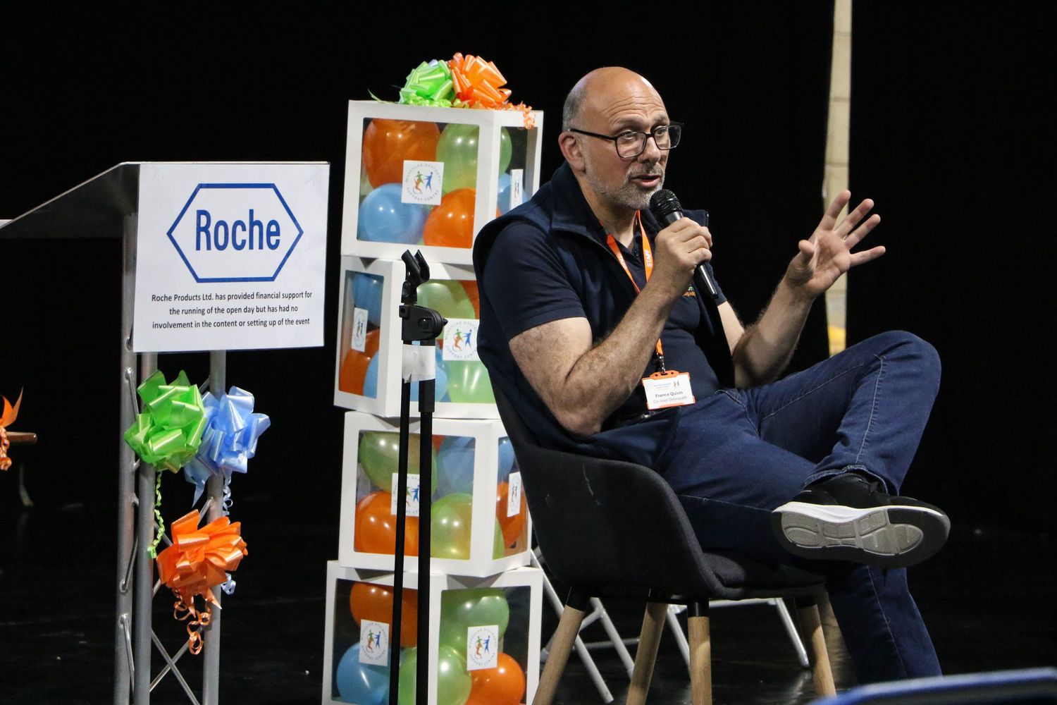 A person in a black shirt sits on stage giving a presentation with colorful decorative elements visible in the background.