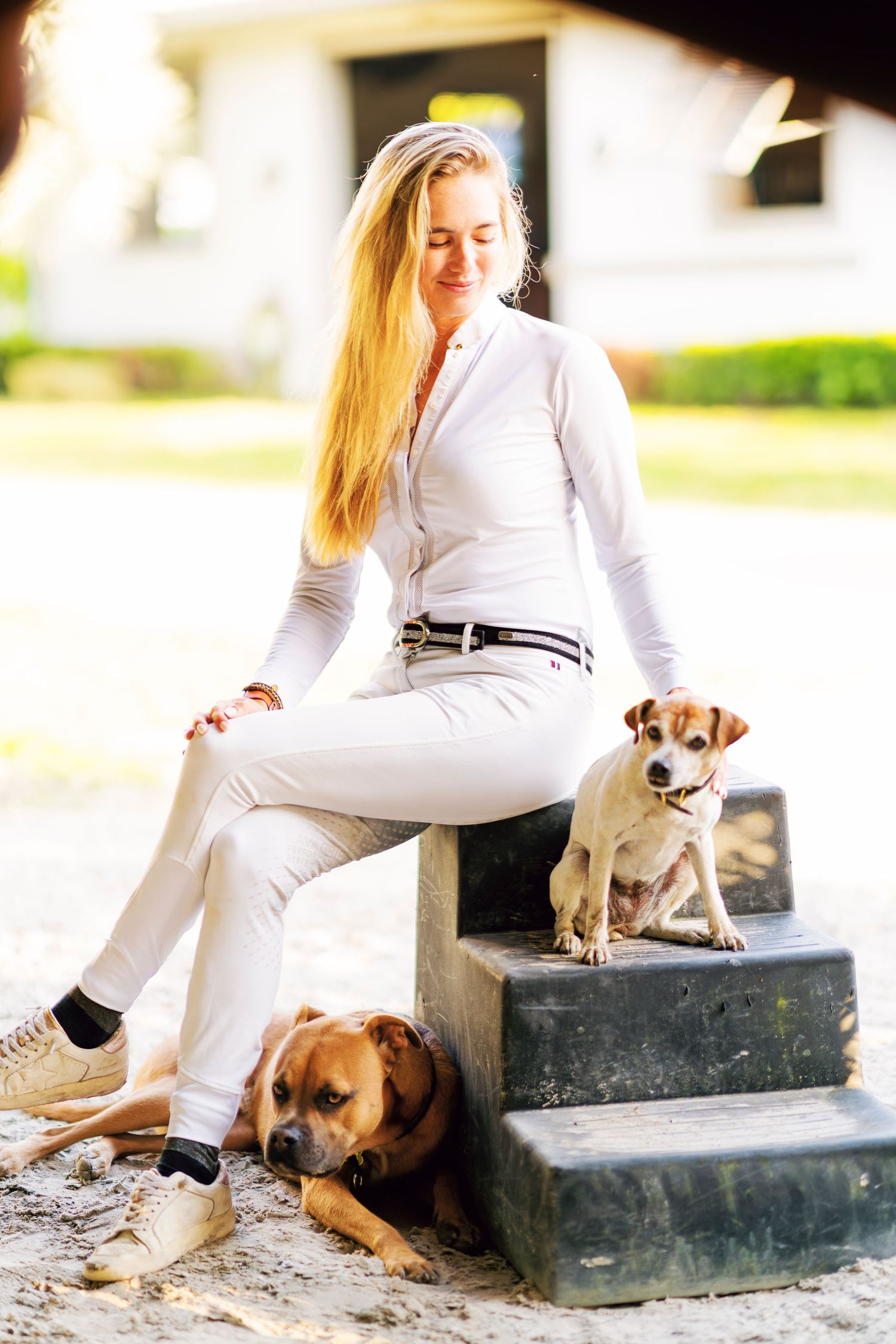 A person in all white attire sits on concrete steps with two small dogs in a sun-drenched outdoor setting.