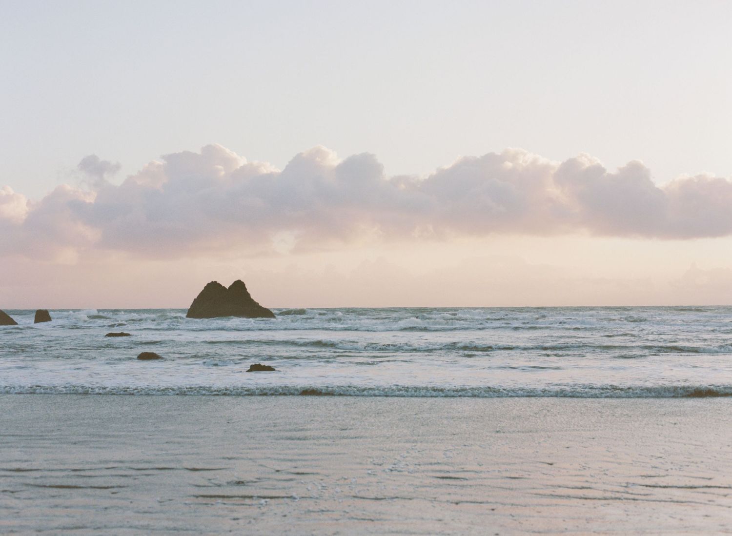 Scenic panoramic view of a beach at sunset with waves crashing against rocky outcrops in the distance.