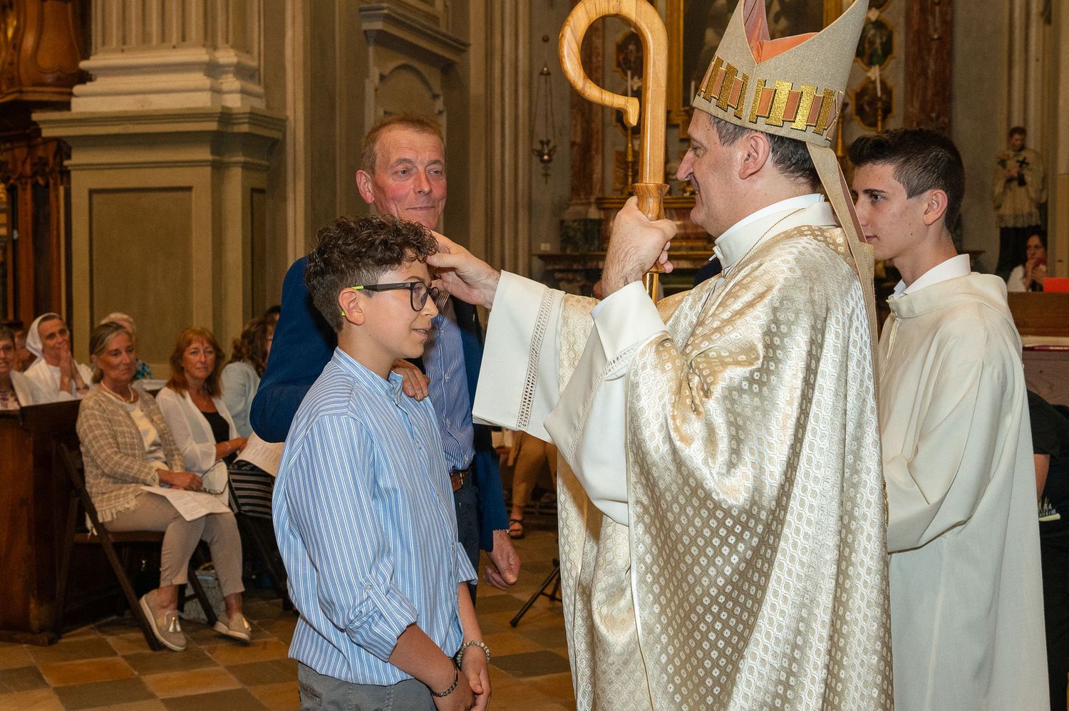Religious confirmation ceremony taking place inside ornate church with bishop performing blessing.