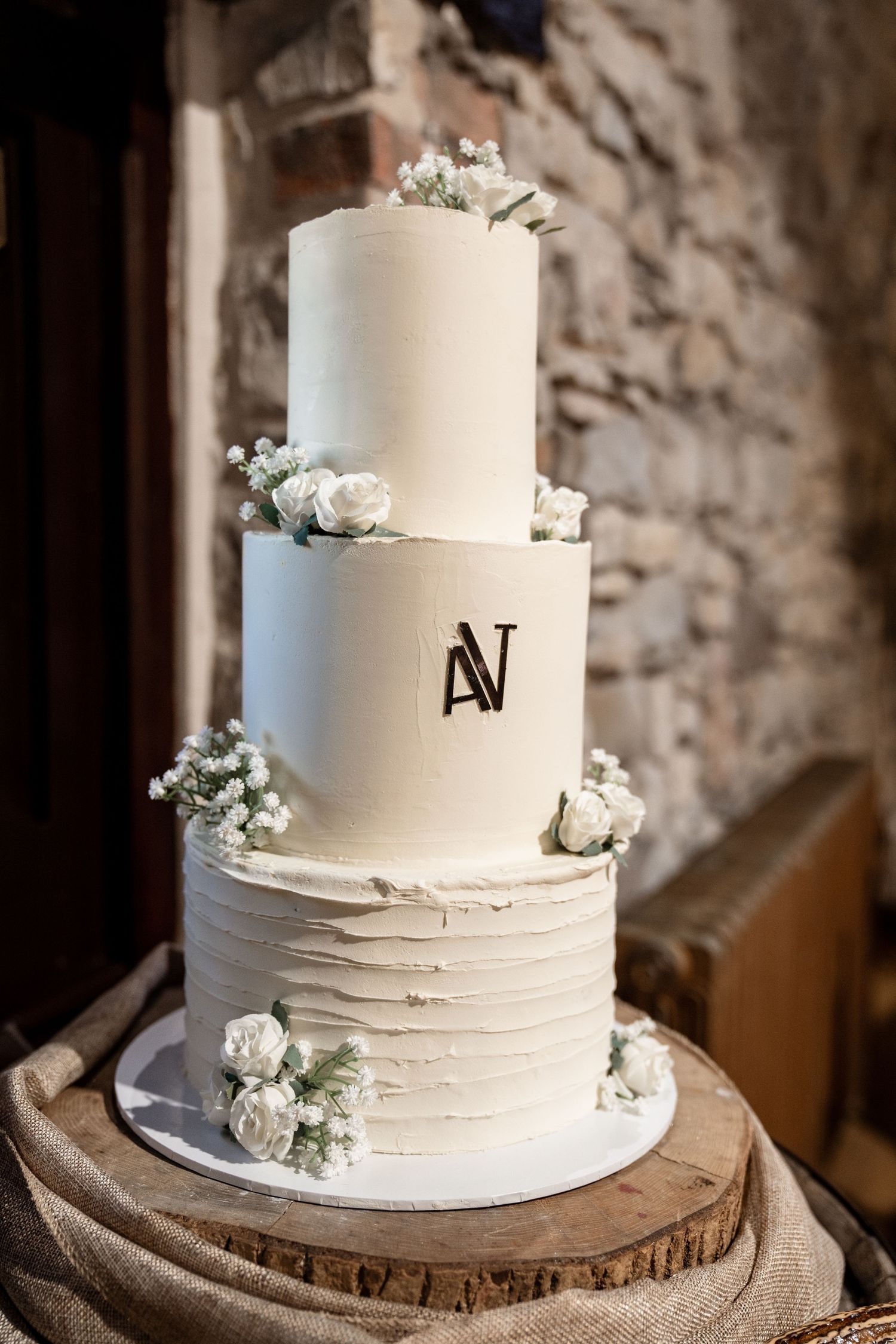 Elegant three-tiered white wedding cake with monogram and baby's breath flowers displayed on rustic wood slice.