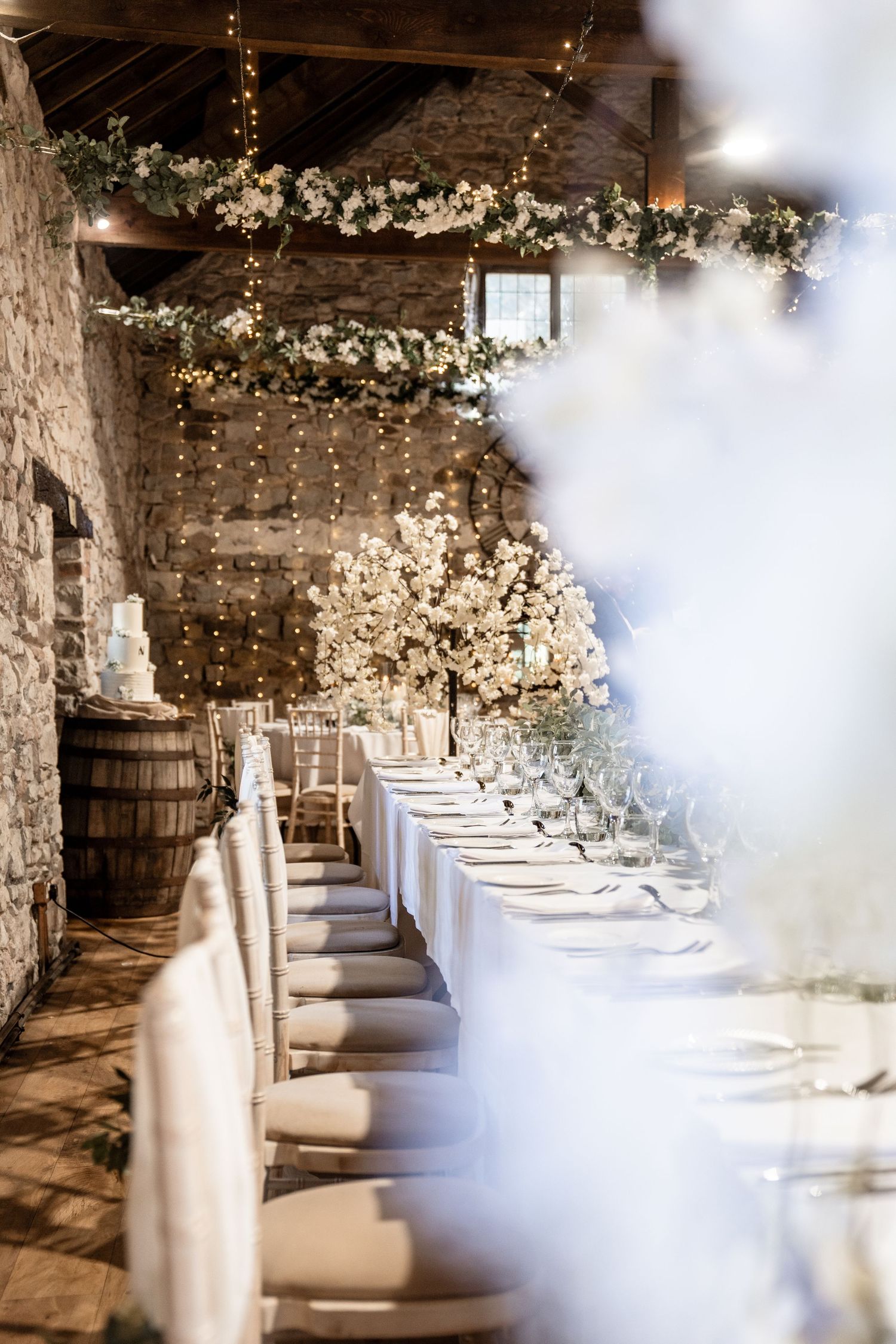 Rustic barn wedding reception setup with white tablecloths, string lights and floral garlands decorating stone walls.