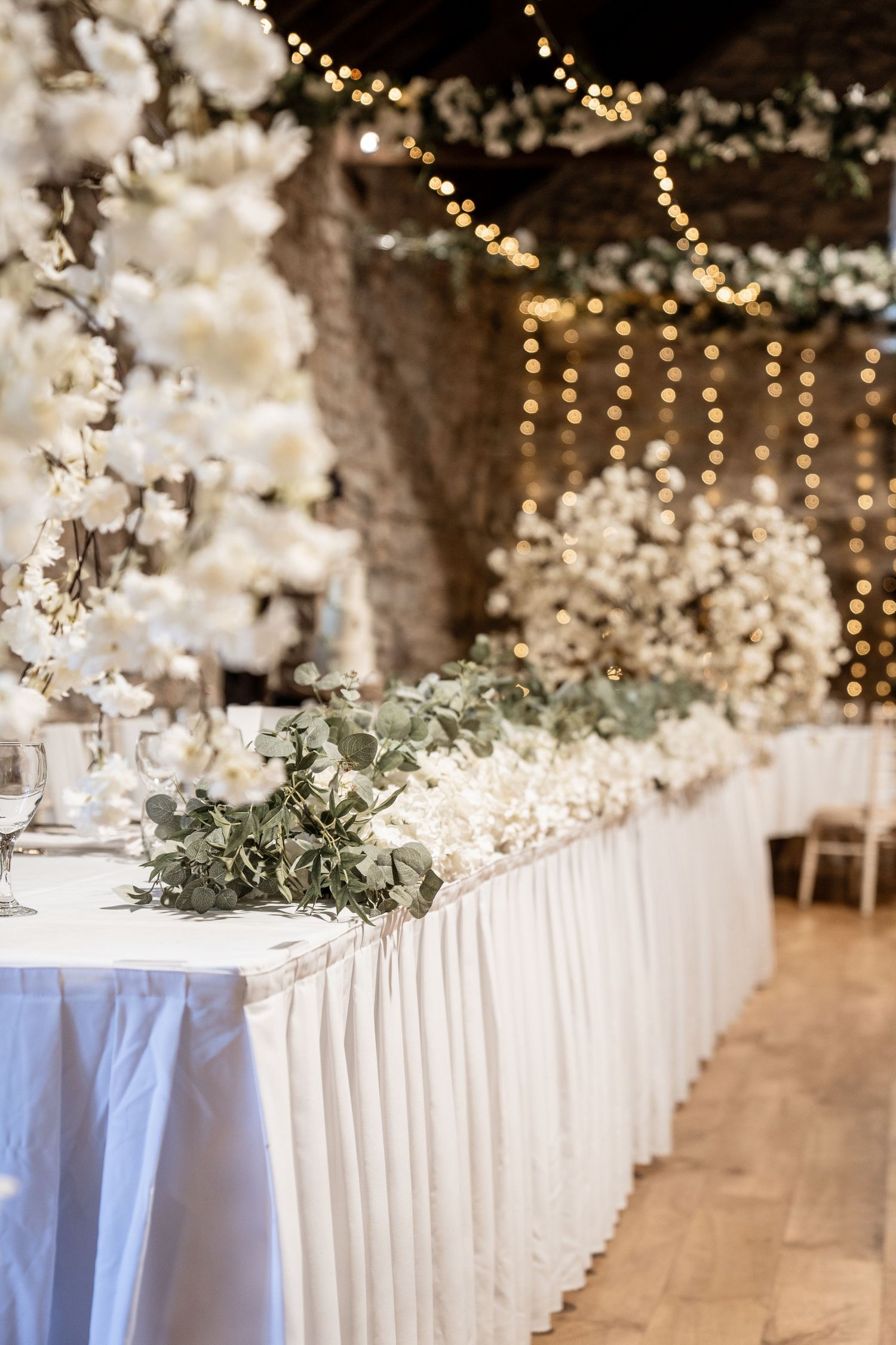 White tablecloths and greenery decorate a long reception table under twinkling lights at an elegant wedding venue.