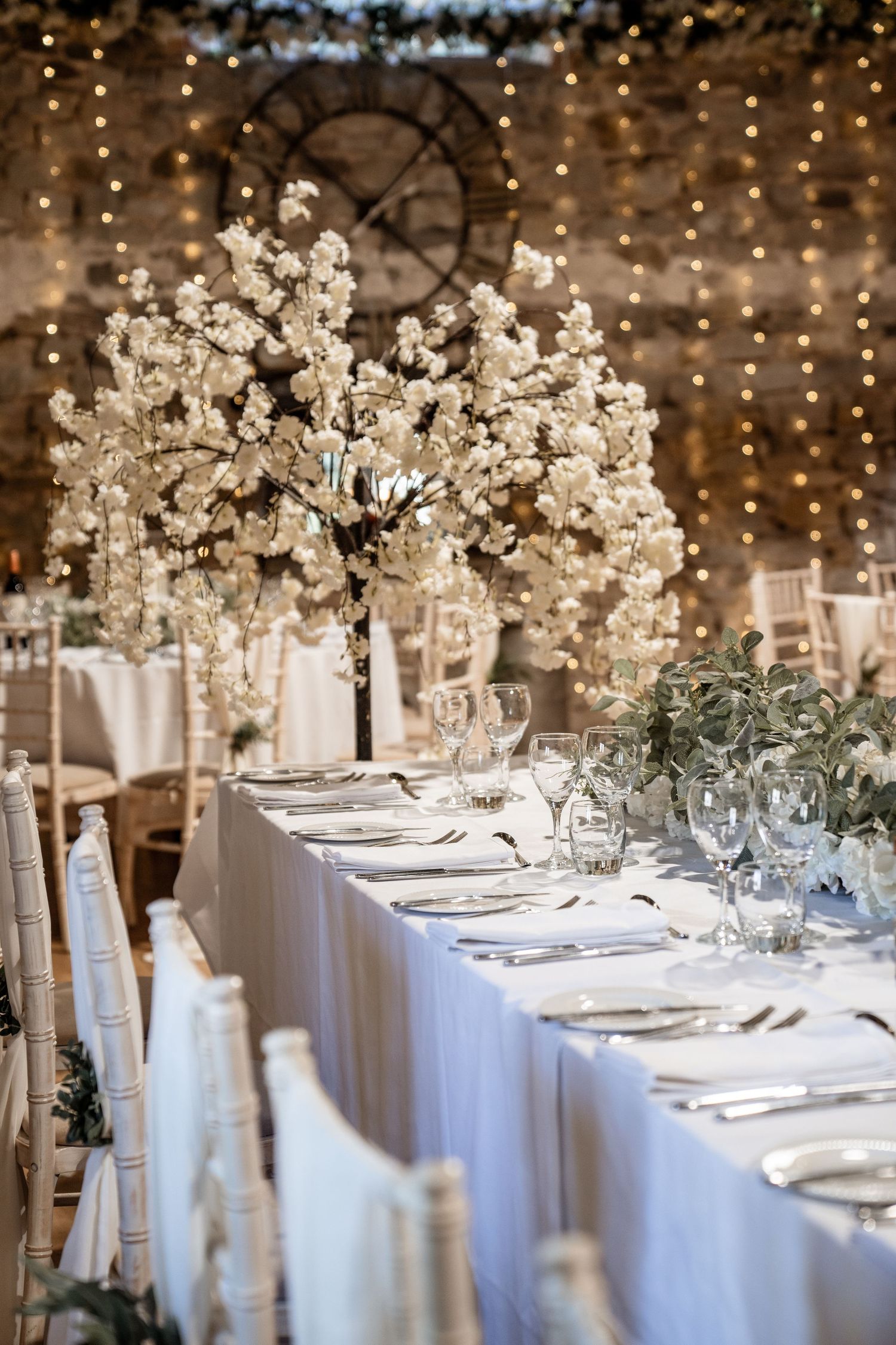 Elegant wedding reception tables with white linens and tall floral centerpieces under twinkling string lights.
