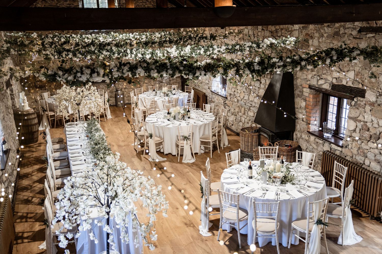 Elegant wedding reception venue decorated with white linens, rustic wooden beams, and twinkling lights overhead.